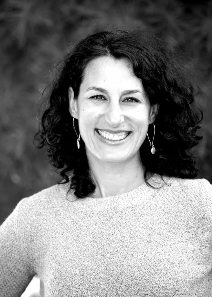 Smiling person with curly hair and earrings, wearing a textured sweater.