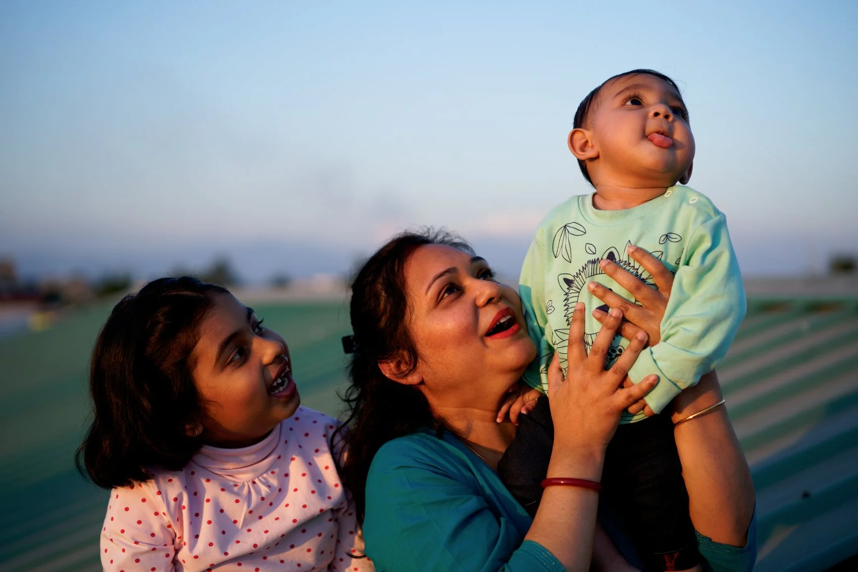 A woman holding a young boy up in the air while a girl looks on happily during sunset outdoors.