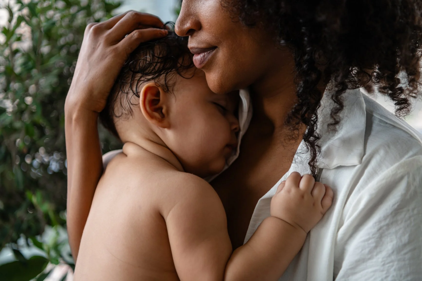A woman tenderly embraces and kisses a young boy, who is resting his head on her shoulder, both with closed eyes, in an outdoor setting with greenery in the background.