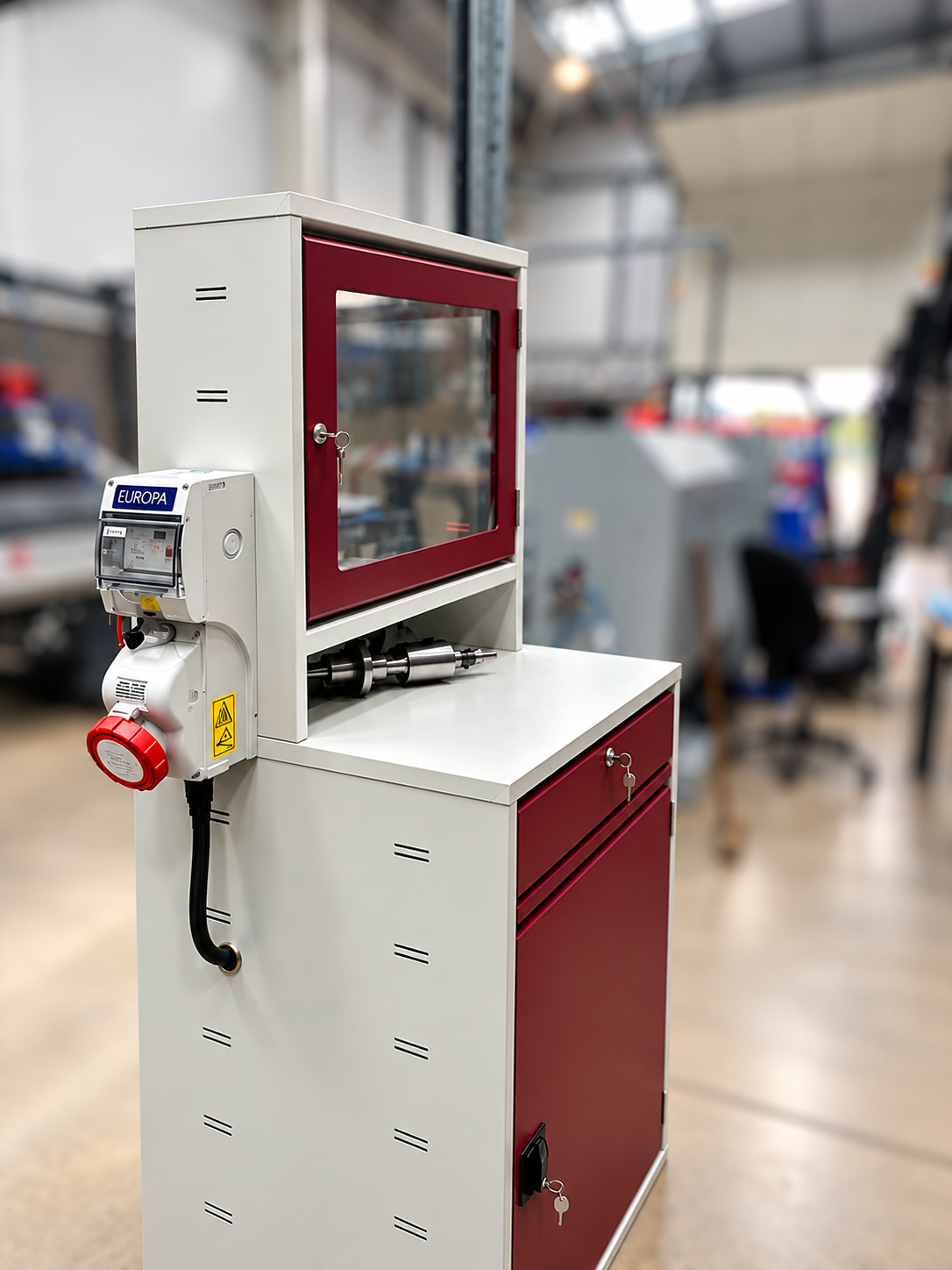A small industrial cabinet with a glass window, a keypad, and a gauge mounted on the side, placed on a table in a factory setting.
