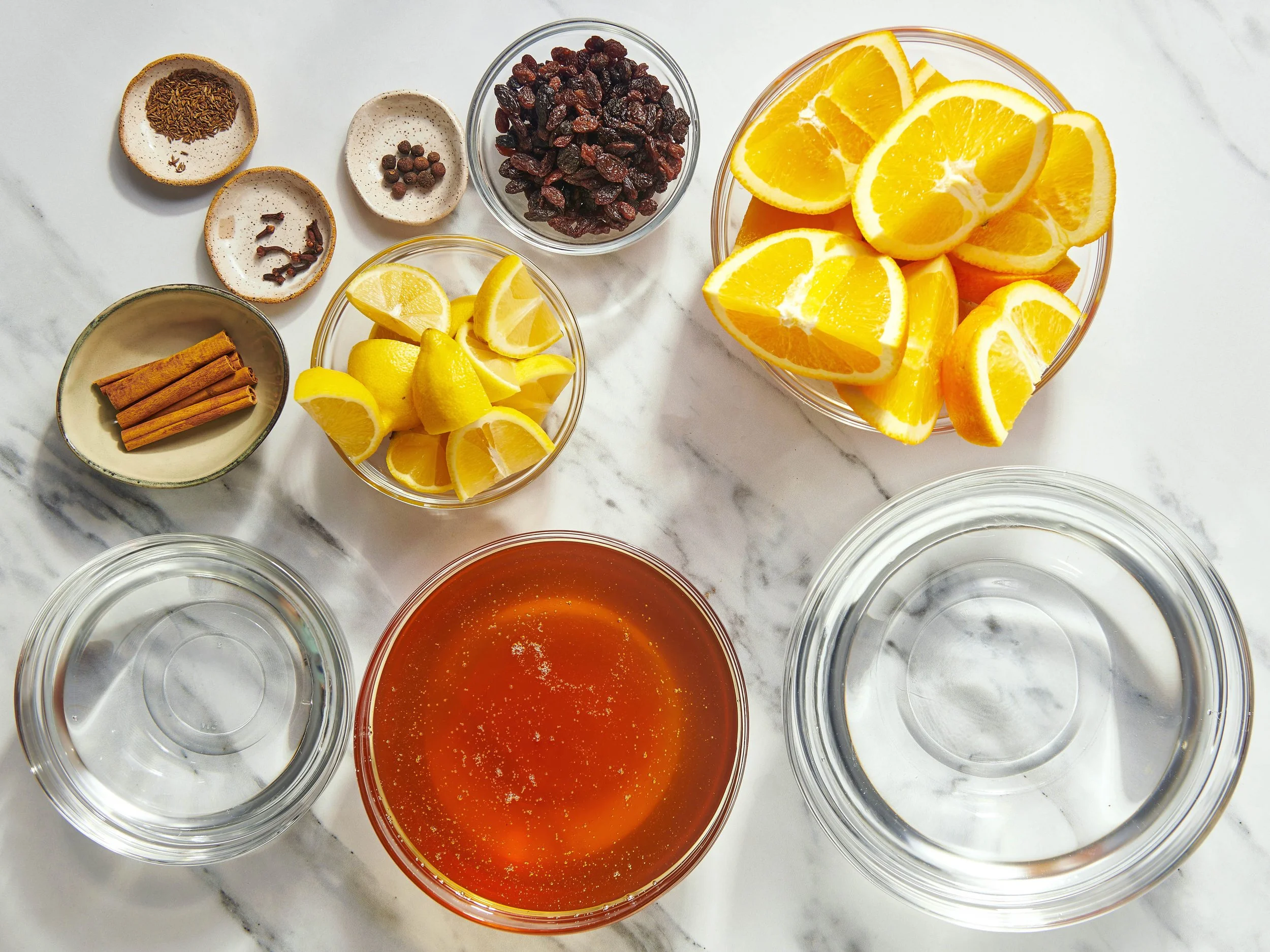 Various bowls and jars containing ingredients such as orange slices, dried cranberries, cinnamon sticks, star anise, allspice berries, and caraway seeds arranged on a white marble surface.