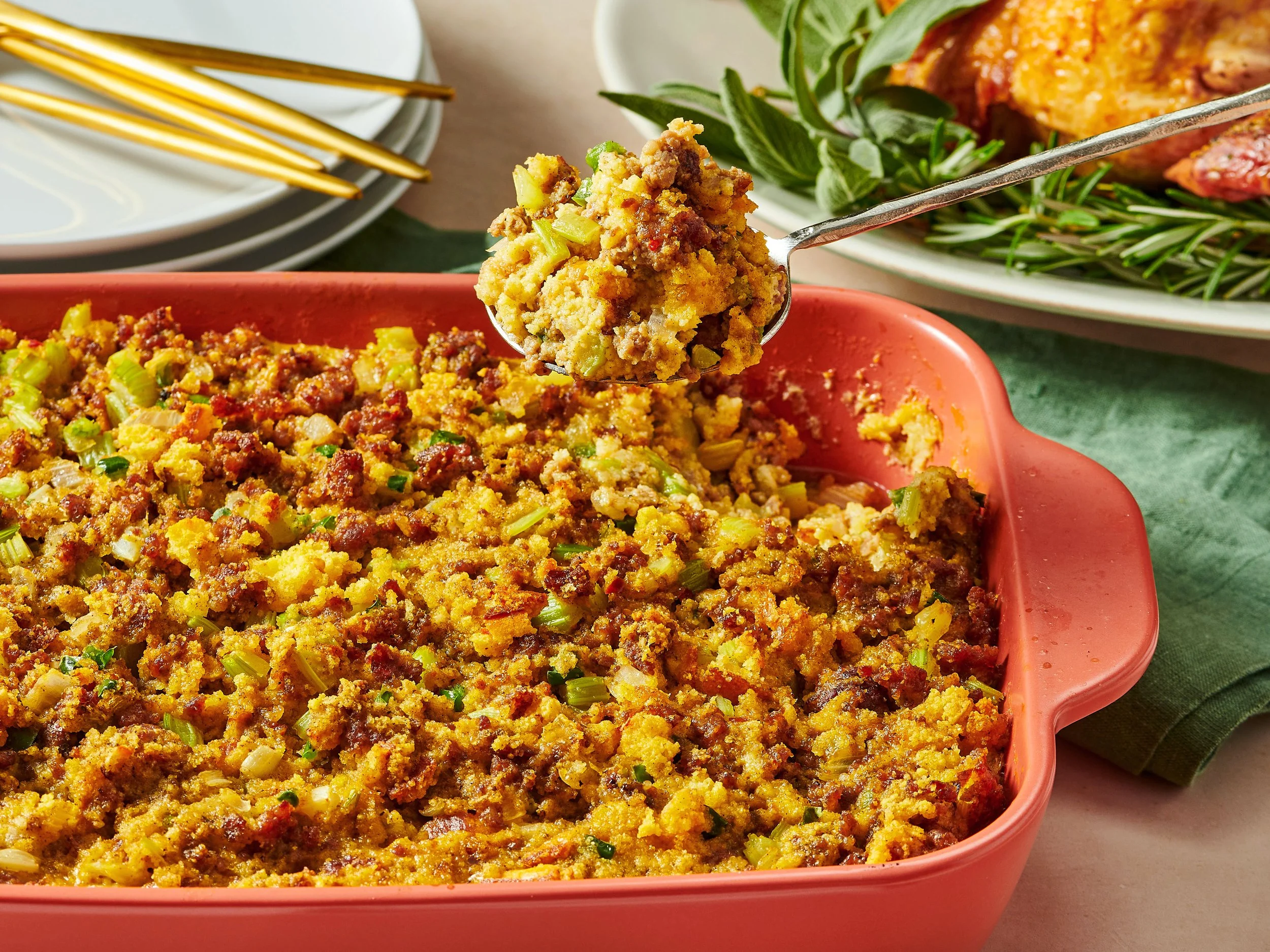A scoop of cooked stuffing in a silver spoon held above a baking dish filled with stuffing. The stuffing contains bread crumbs, celery, and ground meat, surrounded by green herbs. In the background, there are stacked white plates with gold utensils a