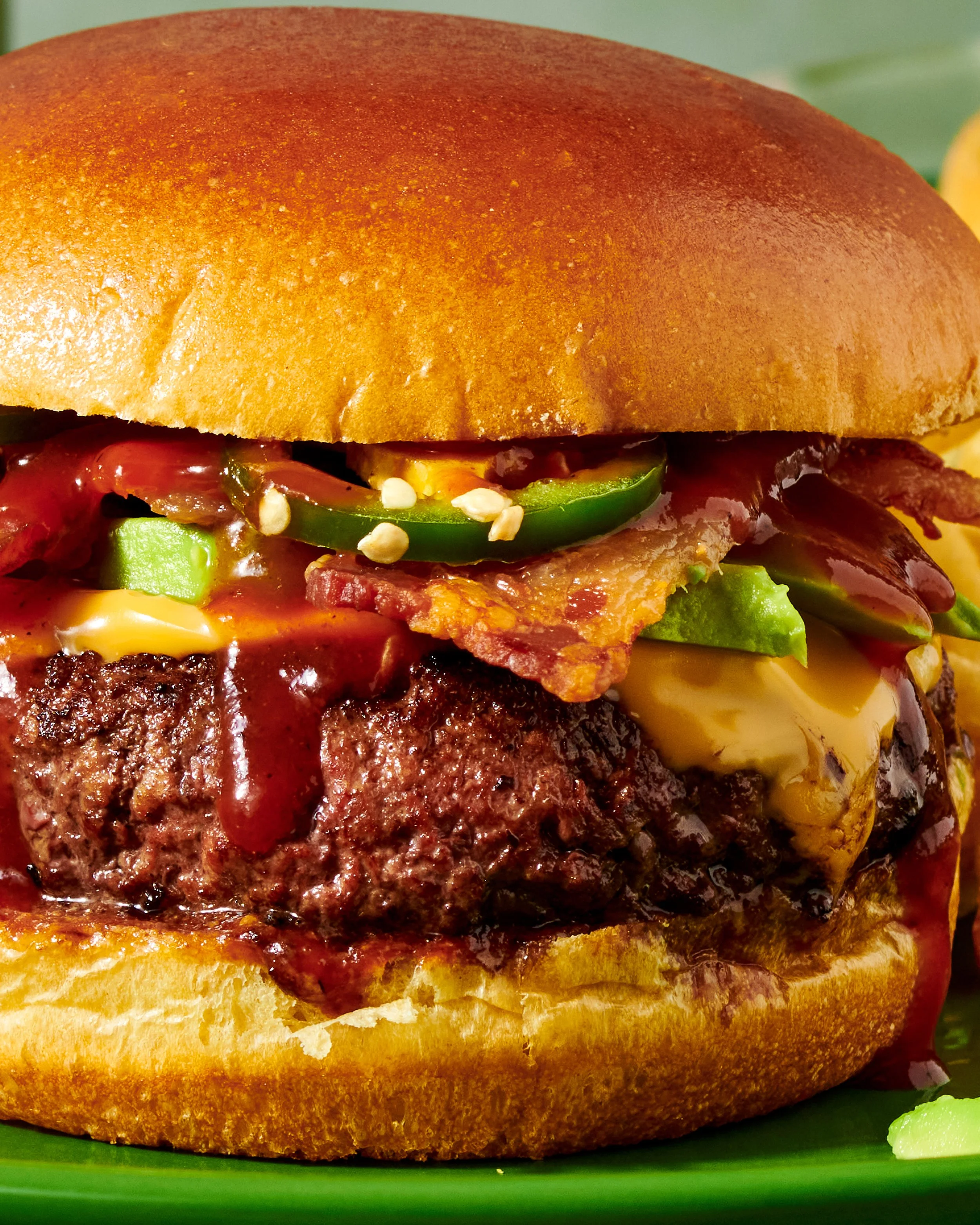 Close-up of a cheeseburger with a bun, cheese, beef patty, bacon, lettuce, tomato, and pickles on a green plate