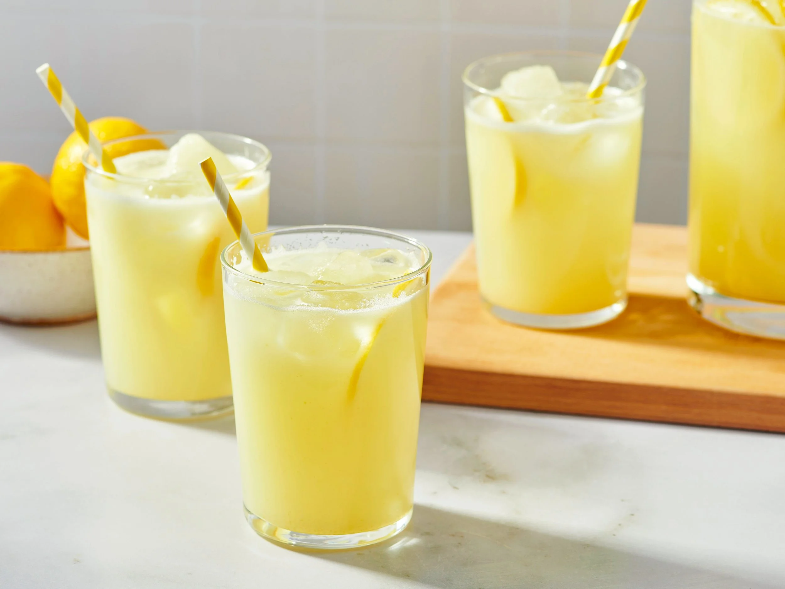 Three glasses of lemonade with ice and yellow and white striped straws on a wooden board, with lemons in the background.