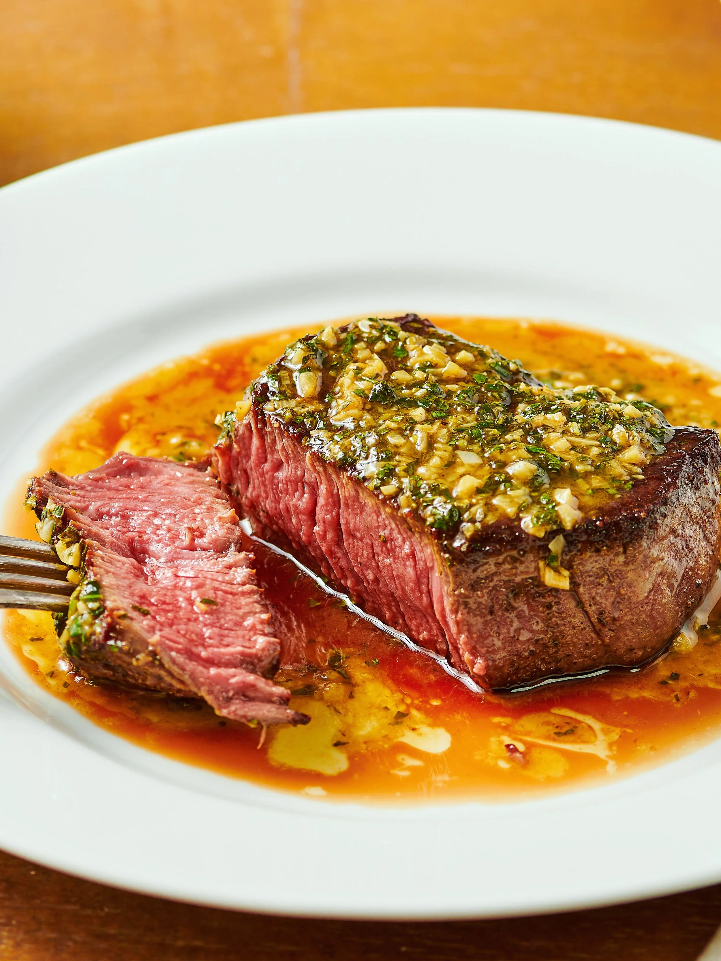A cut of medium-rare steak garnished with chopped garlic and herbs, served with a savory sauce on a white plate.