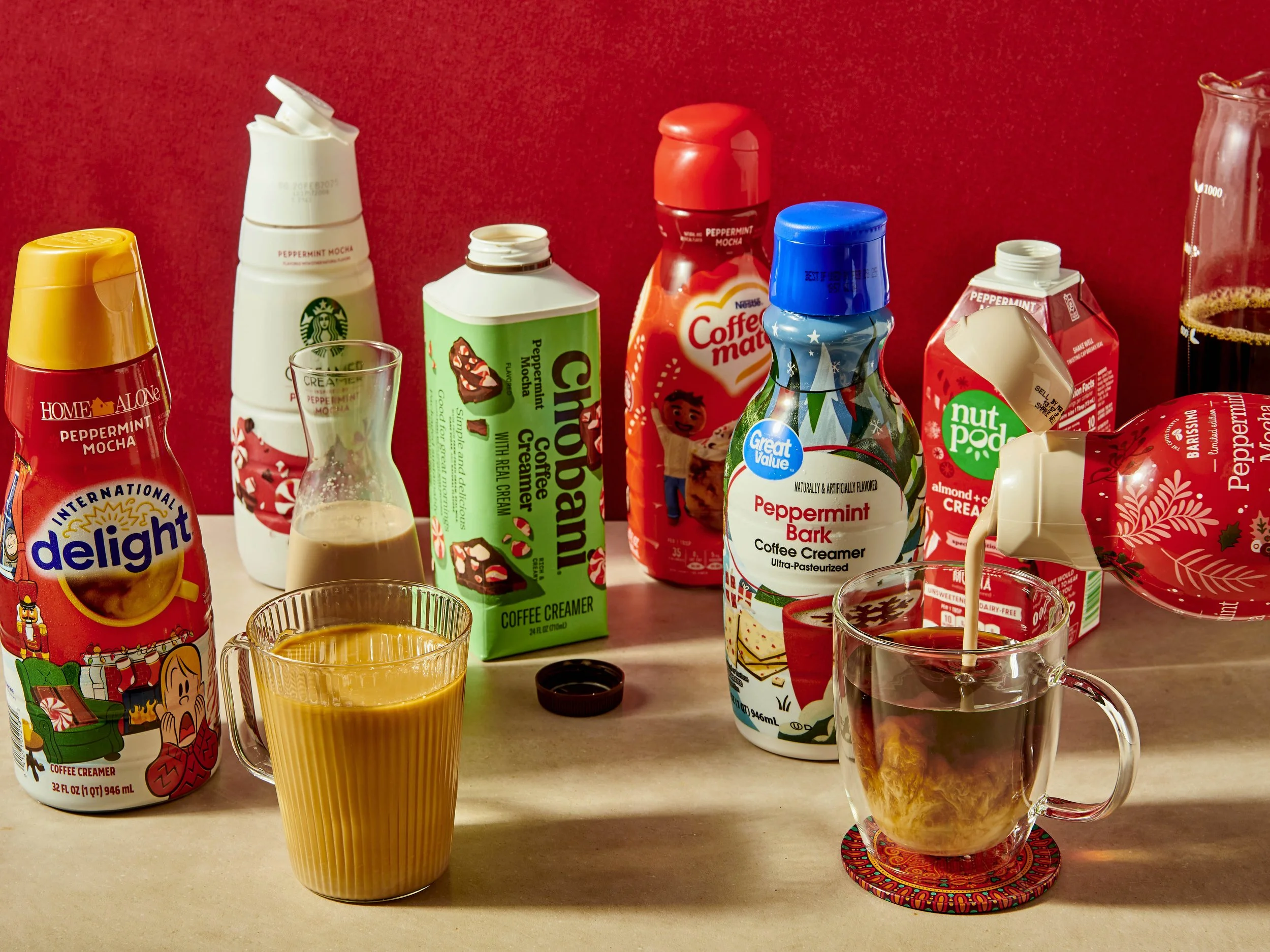 Various coffee creamers, a glass of coffee with cream, and a glass of black coffee on a beige countertop against a red background.