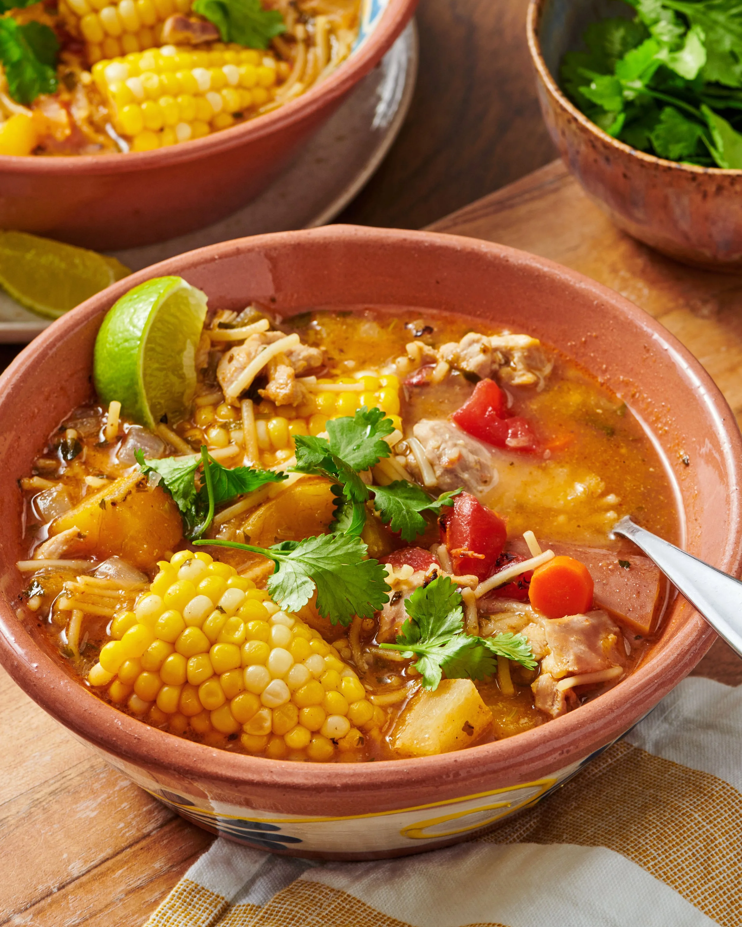 A bowl of chicken tortilla soup garnished with cilantro, lime wedges, corn, shredded cheese, and diced tomatoes, on a wooden table with a folded yellow and white cloth.