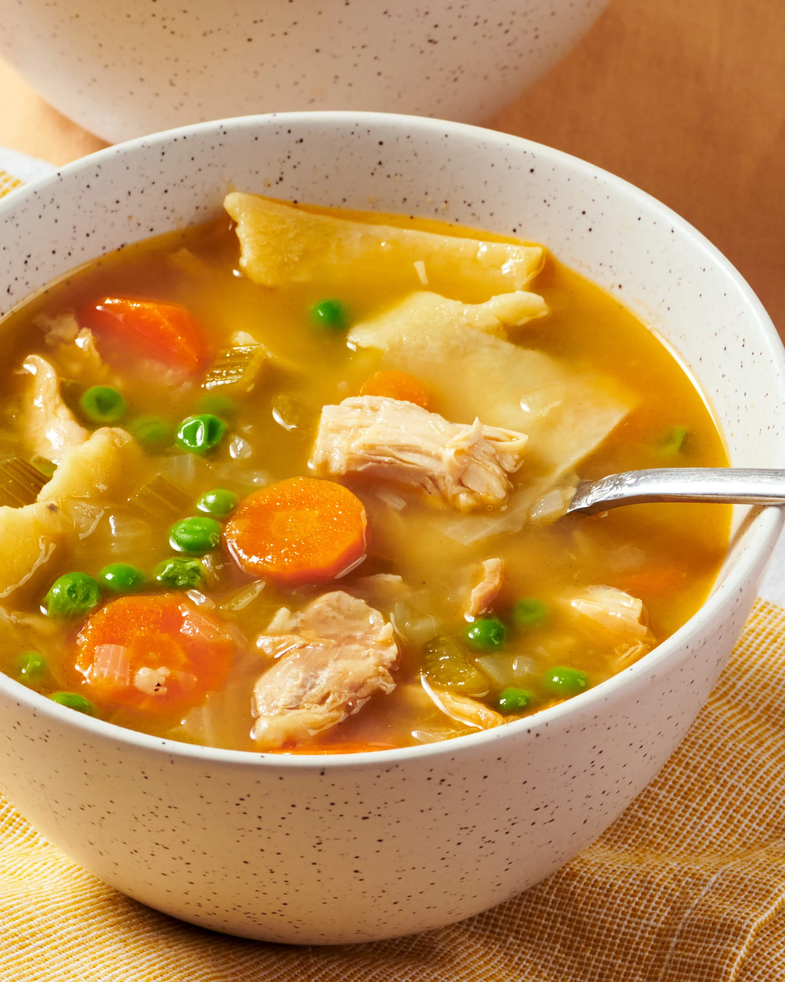 Close-up of a bowl of chicken vegetable soup with carrots, peas, and chicken in a speckled white bowl on a yellow felt placemat.