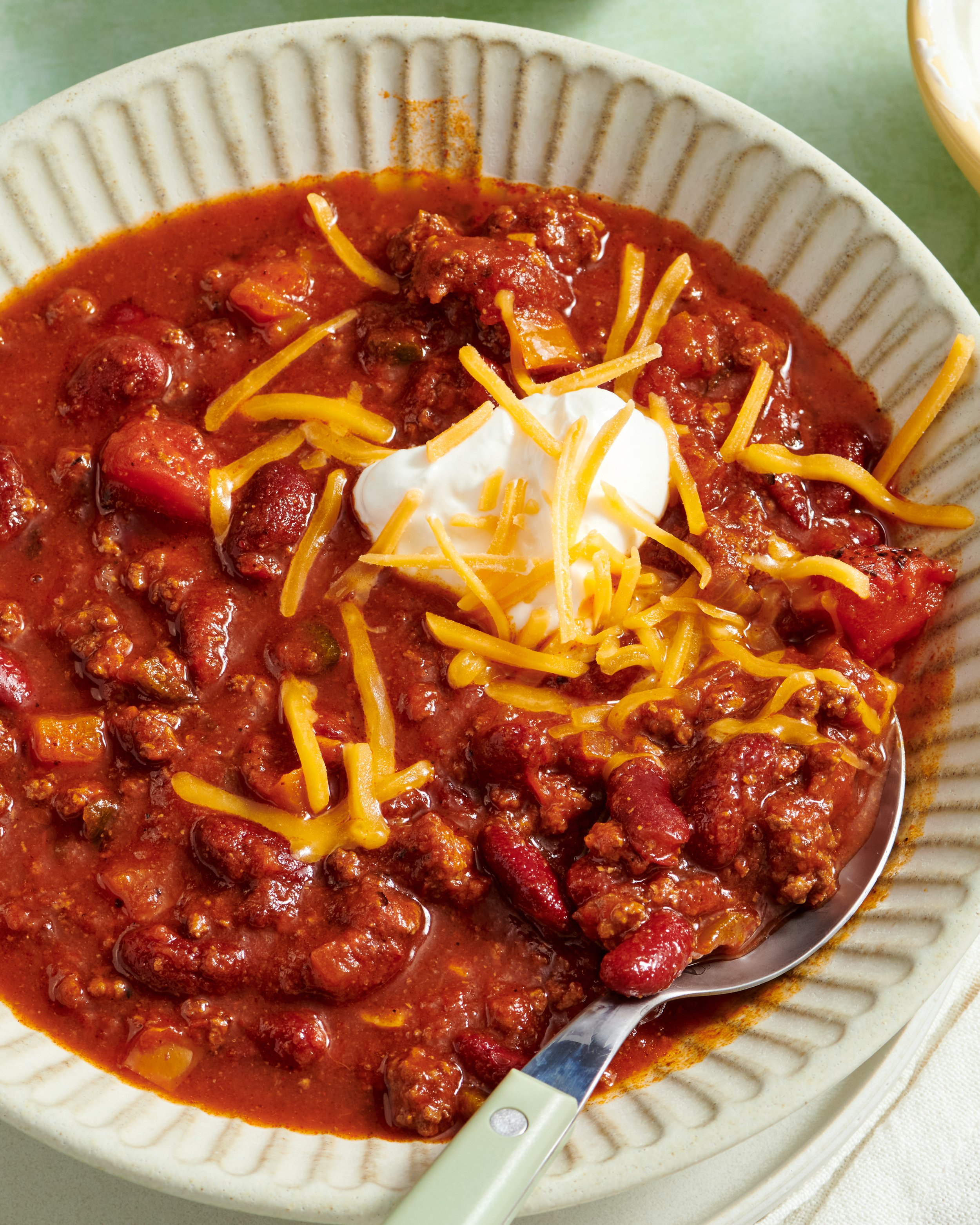 Bowl of chili topped with shredded cheese and sour cream, with a spoon resting in the bowl, on a white cloth.