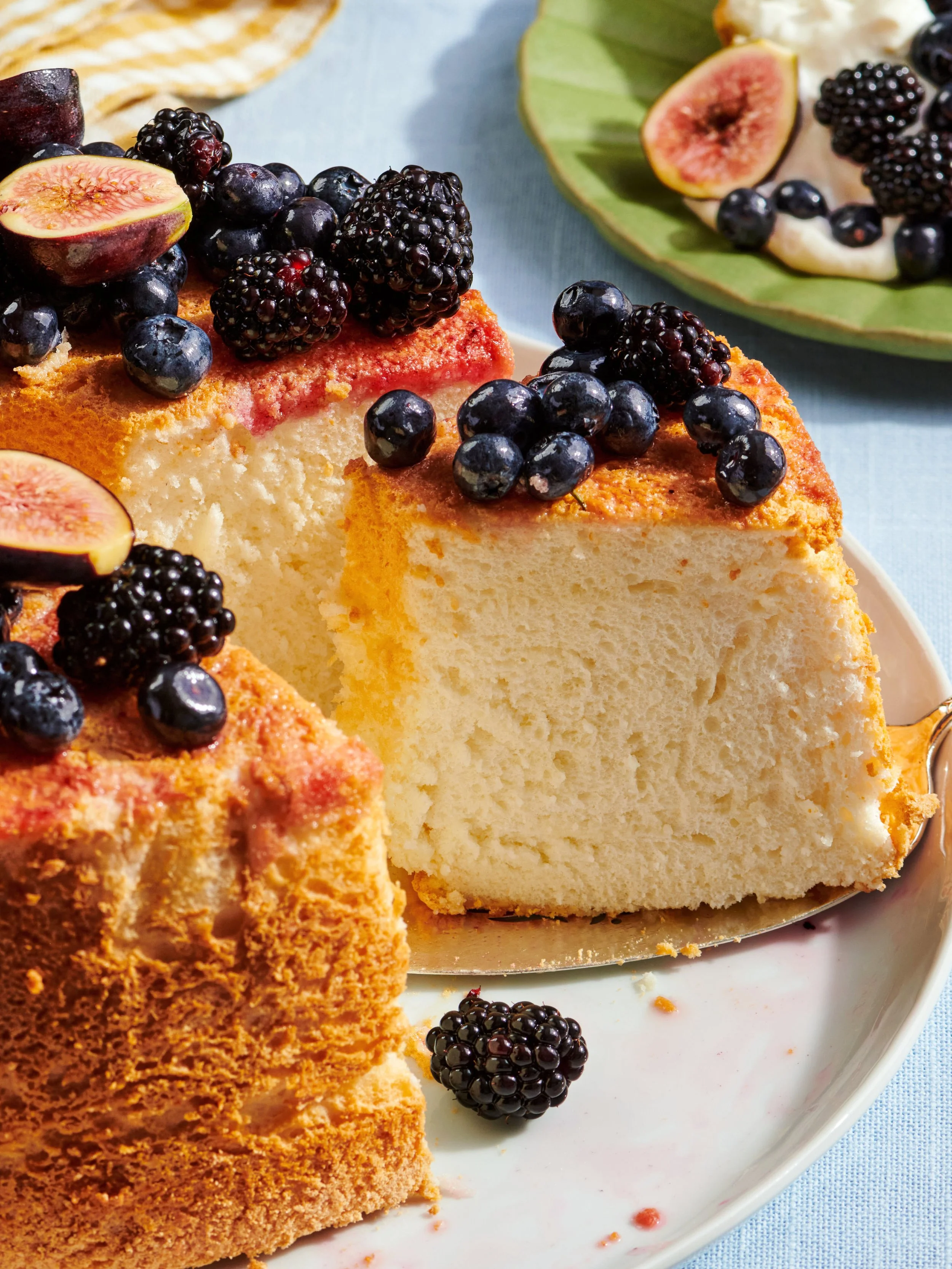 A slice of vanilla bundt cake topped with blackberries, blueberries, and halved figs on a white plate, with more berries and fig slices on a green plate nearby.