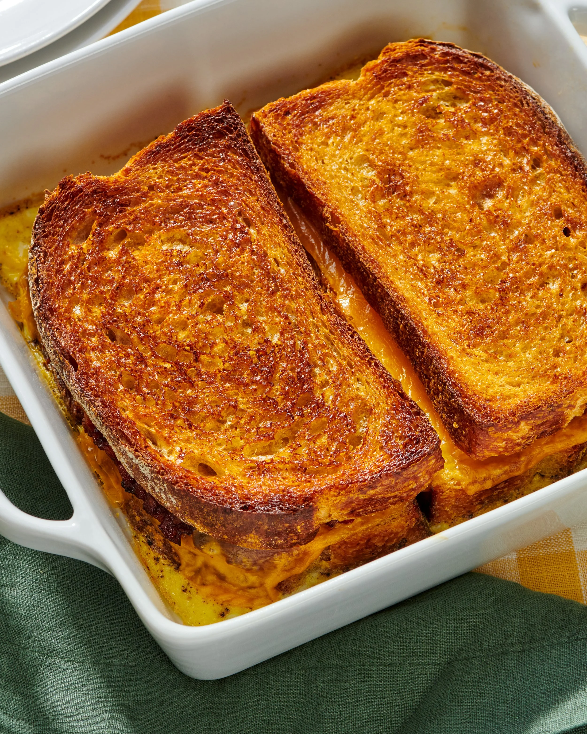 Two slices of French toast with golden-brown crust in a white baking dish, with melted butter underneath, on a dark green cloth.