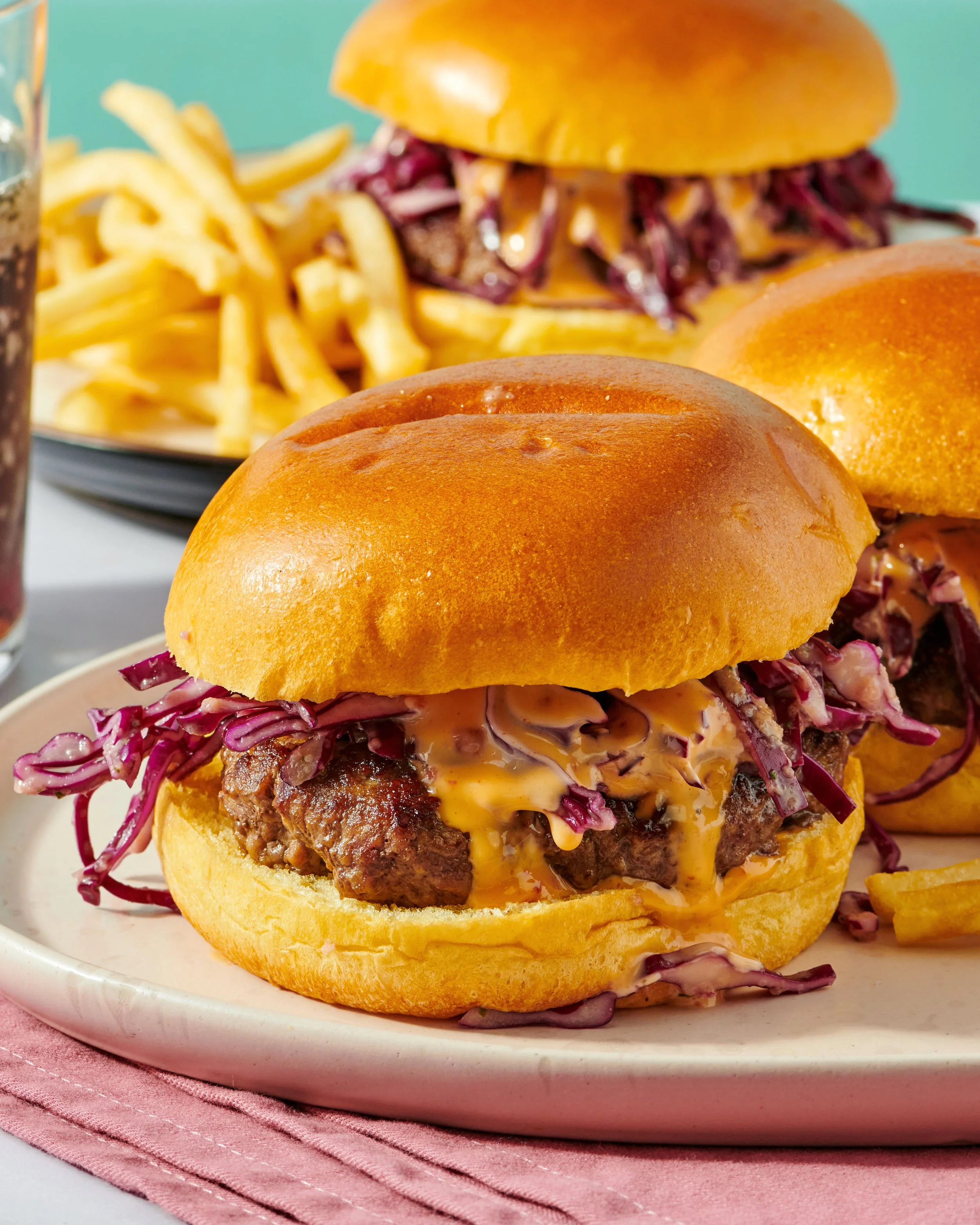 Close-up of two cheeseburgers with coleslaw on soft buns, served on a white plate with a pink napkin underneath.