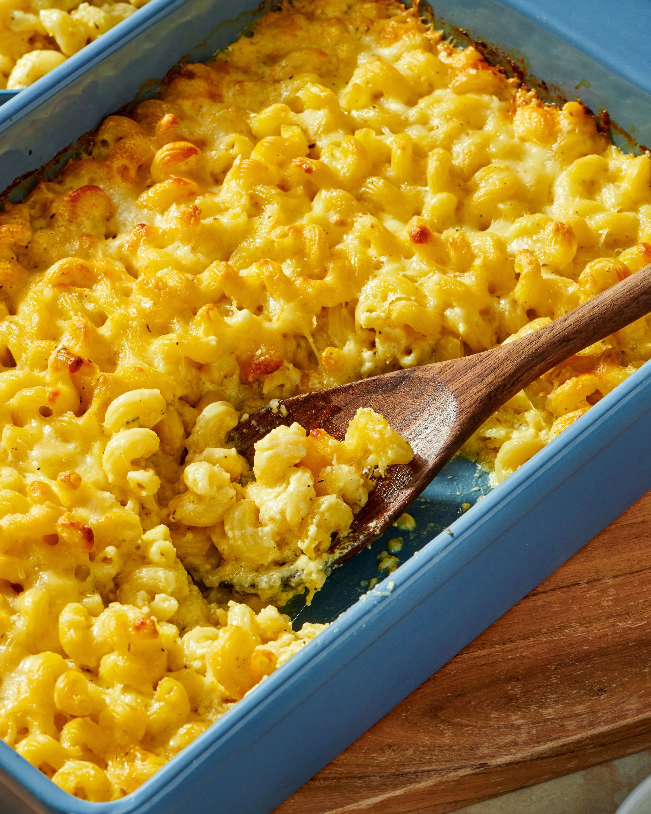 Baked macaroni and cheese in a blue dish with a wooden spoon, with some baked cheese crust on top and pasta visible underneath.