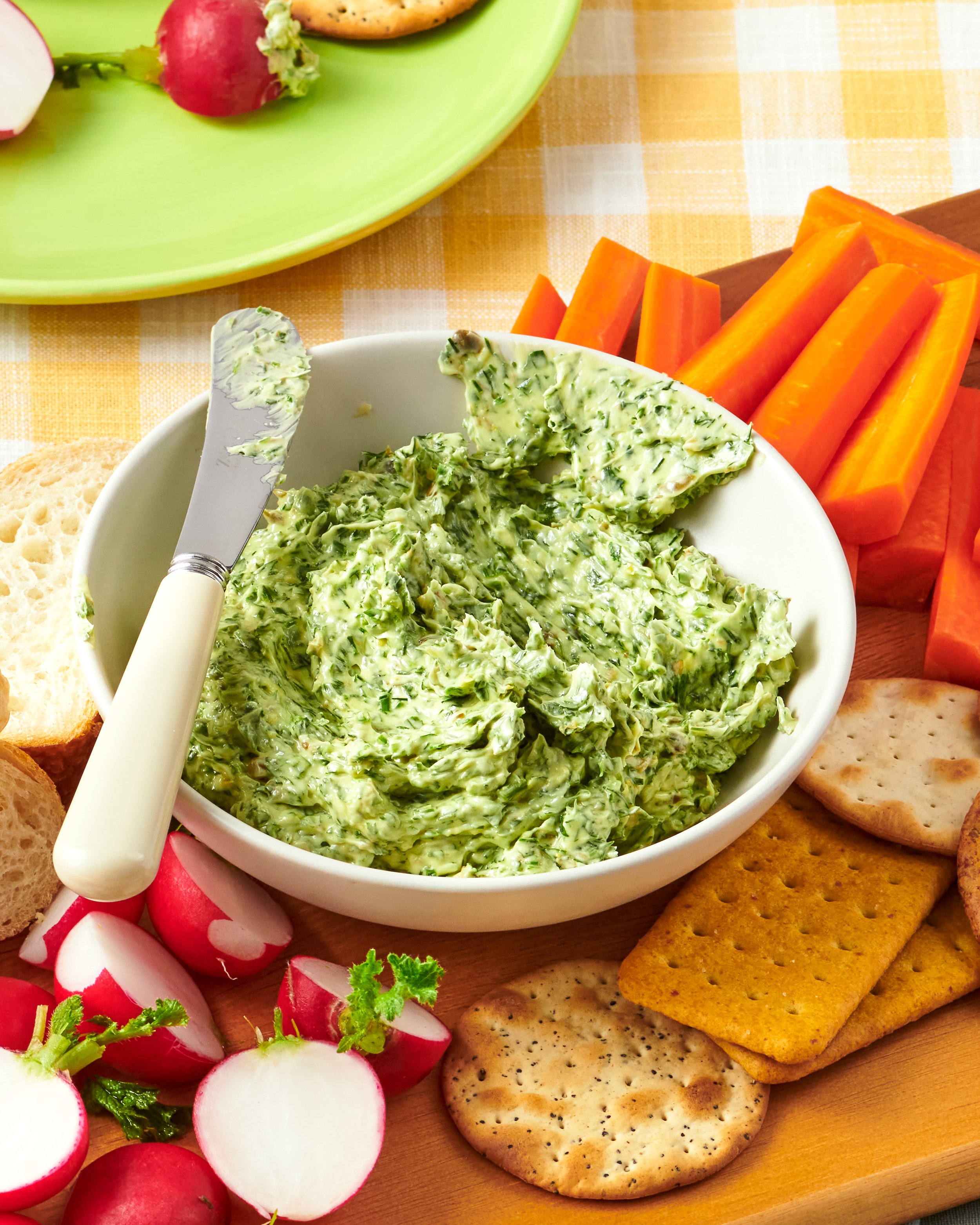 A bowl of creamy spread with herbs and a butter knife, surrounded by sliced radishes, crackers, carrot sticks, and a green plate with other vegetables on a wooden surface.