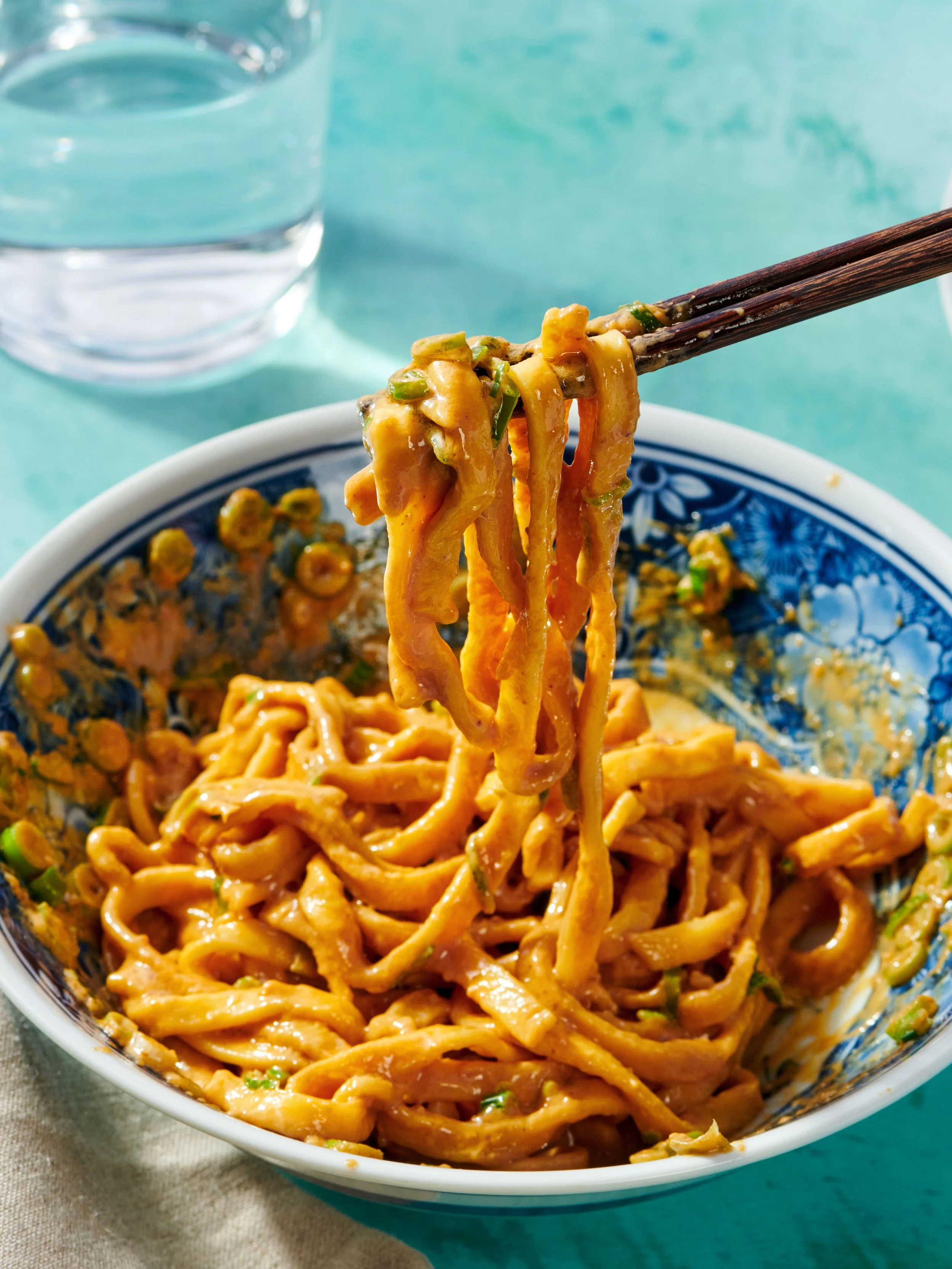 Close-up of a bowl of Asian-style noodles with sauce, garnished with chopped green onions, and chopsticks lifting some noodles. A glass of water is in the background.