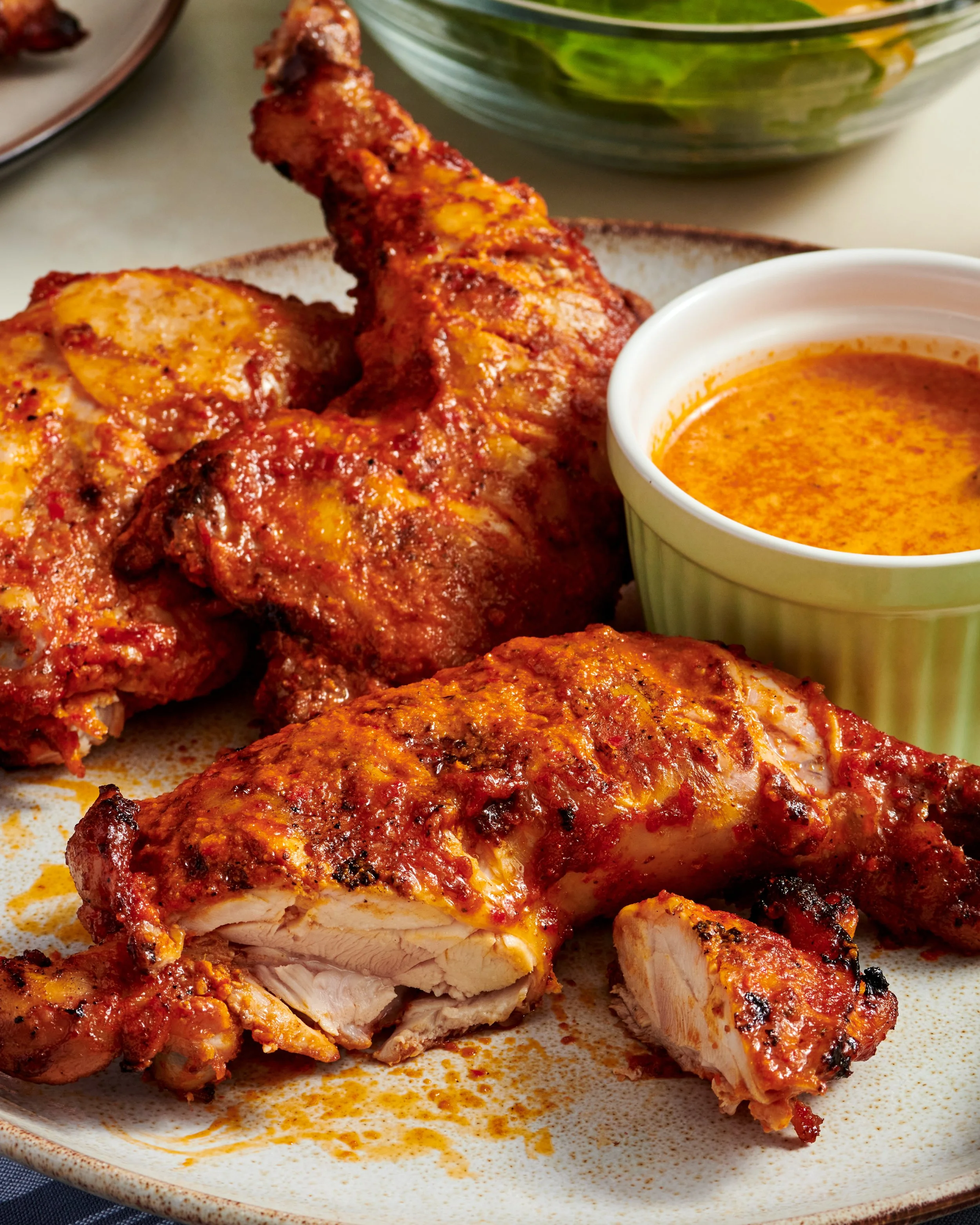 Close-up of baked chicken drumsticks with a reddish seasoning, served on a plate with a small cup of orange-colored sauce or gravy.