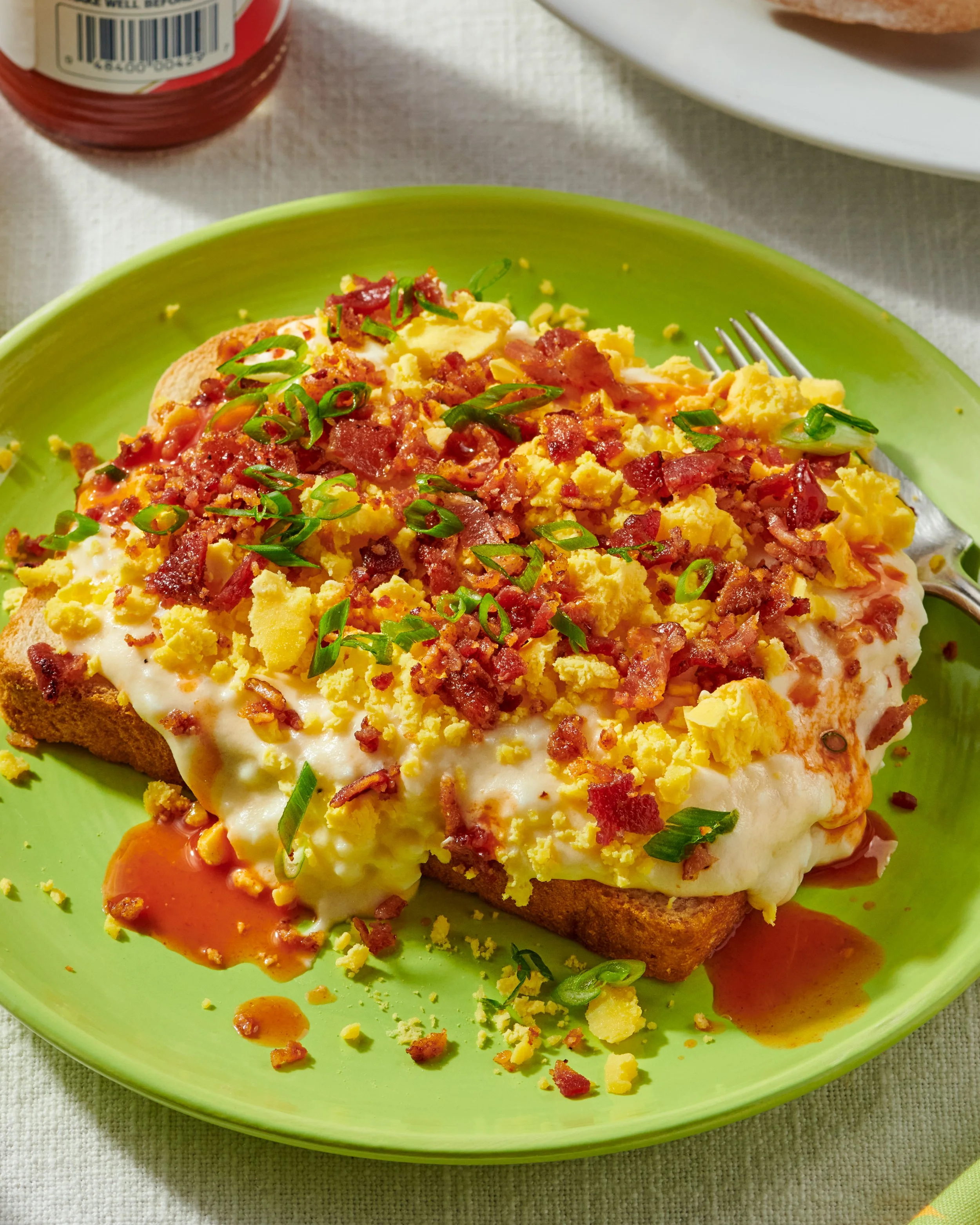 Open-faced breakfast toast topped with scrambled eggs, bacon bits, chopped green onions, and sauce on a green plate.
