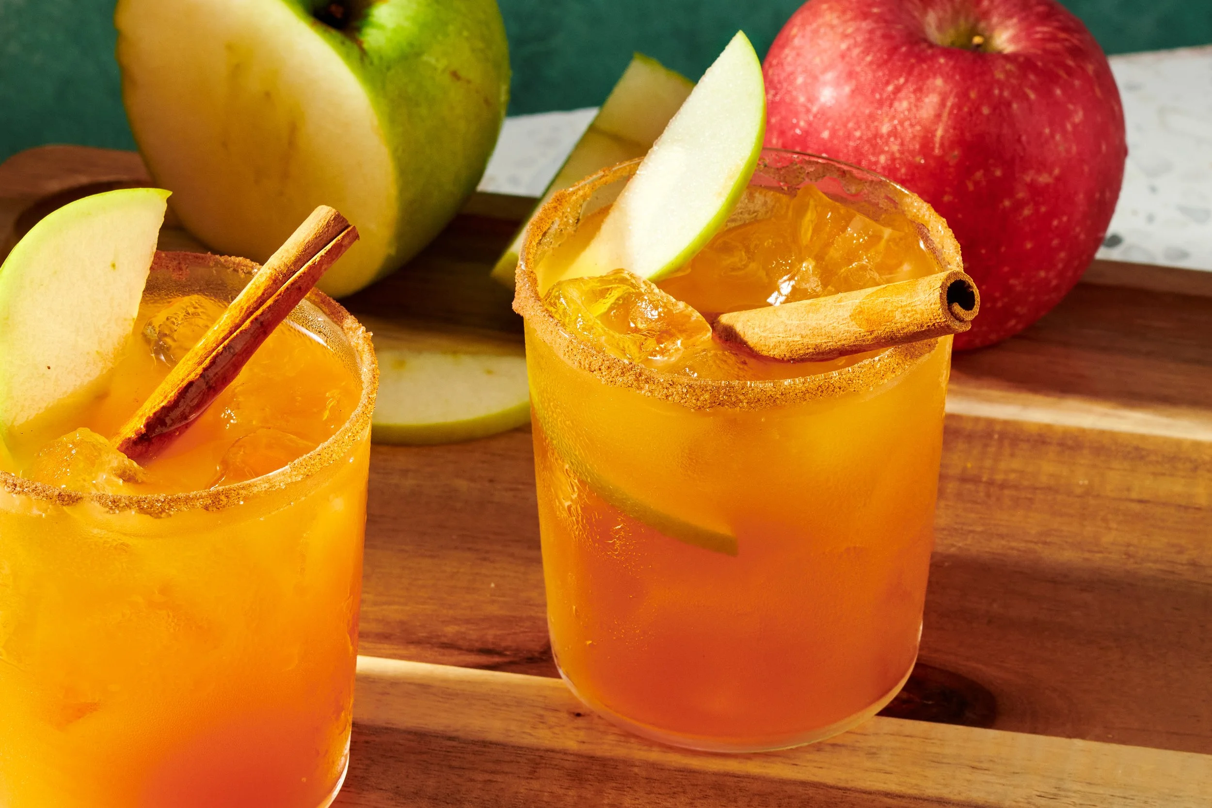 Two glasses of orange-colored beverage garnished with apple slices and cinnamon sticks, with whole green and red apples in the background on a wooden surface.
