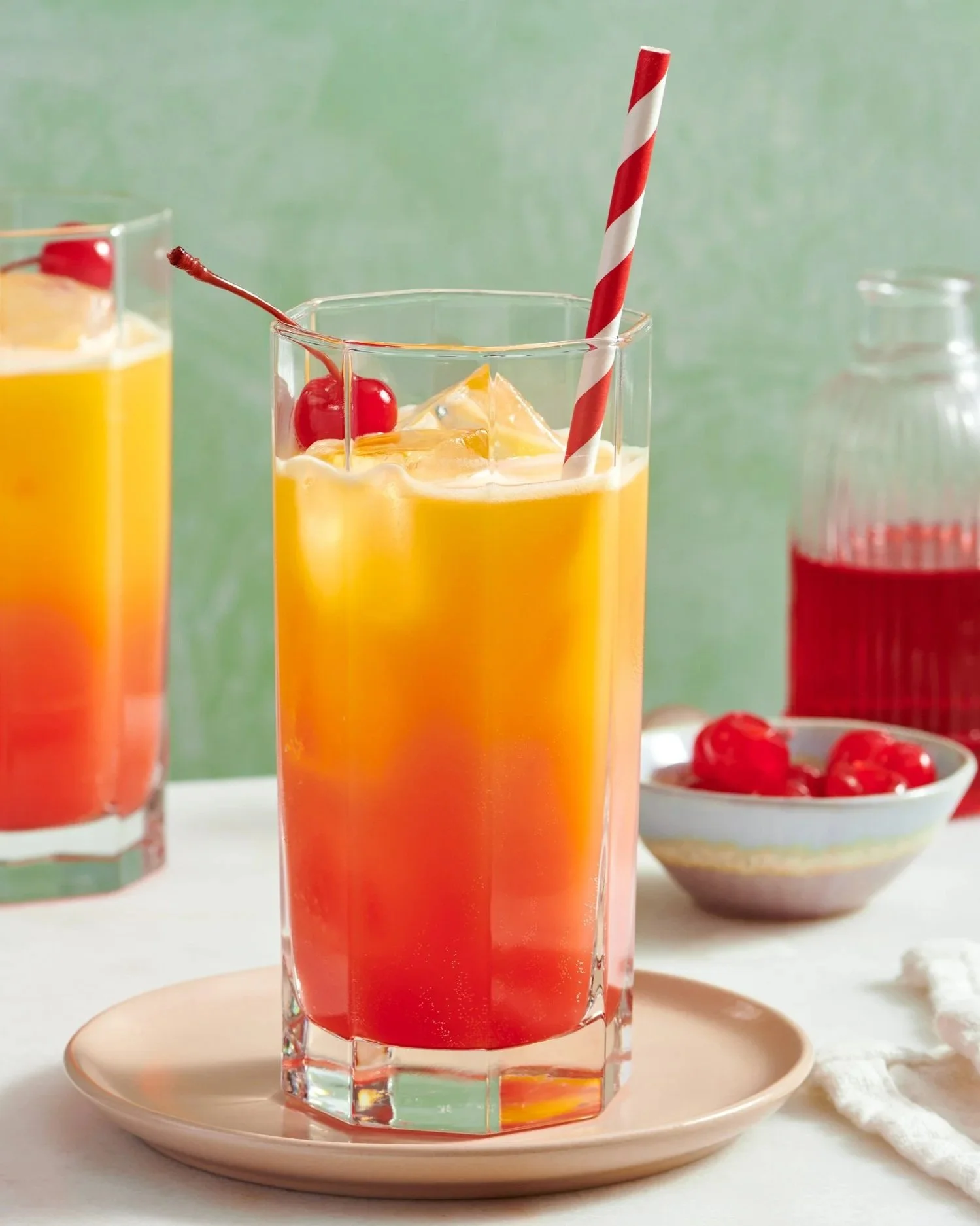 A glass of layered red, orange, and yellow juice with ice cubes, garnished with cherries and a red and white striped straw, on a white plate.
