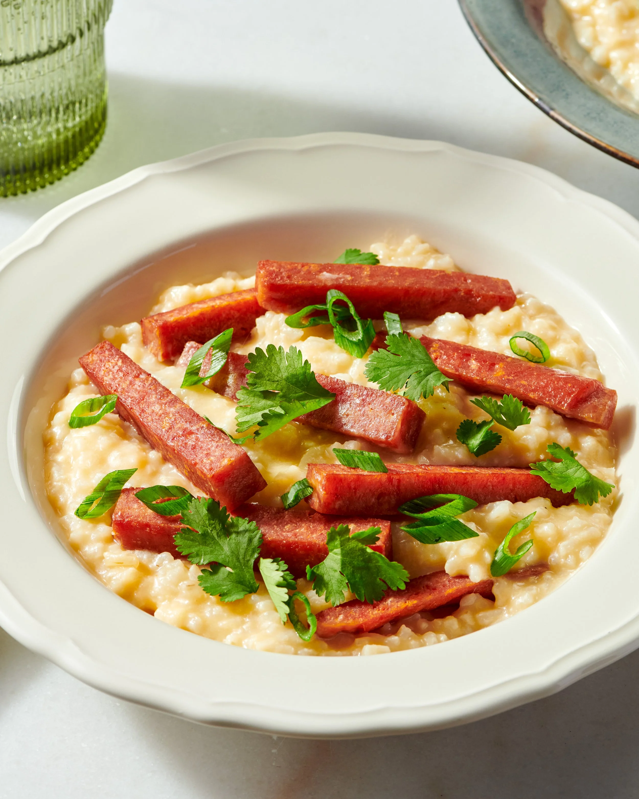 Close-up of a bowl of creamy rice pudding topped with sliced sausage, chopped green onions, and cilantro, on a white surface with a green textured glass and a partially visible dish.