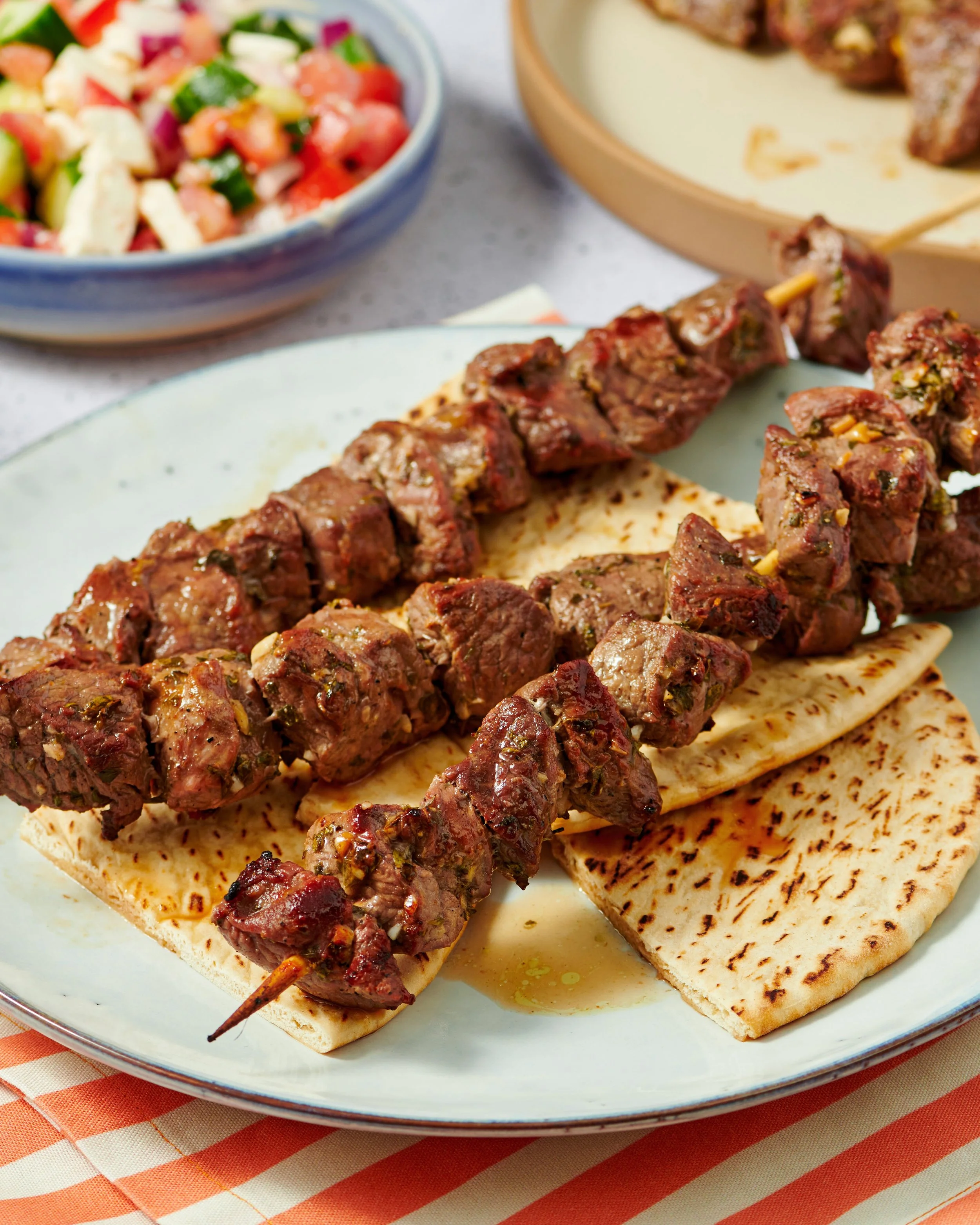 Plate with grilled skewers of beef, pieces of pita bread, and a side bowl of chopped vegetable salad on a striped tablecloth.