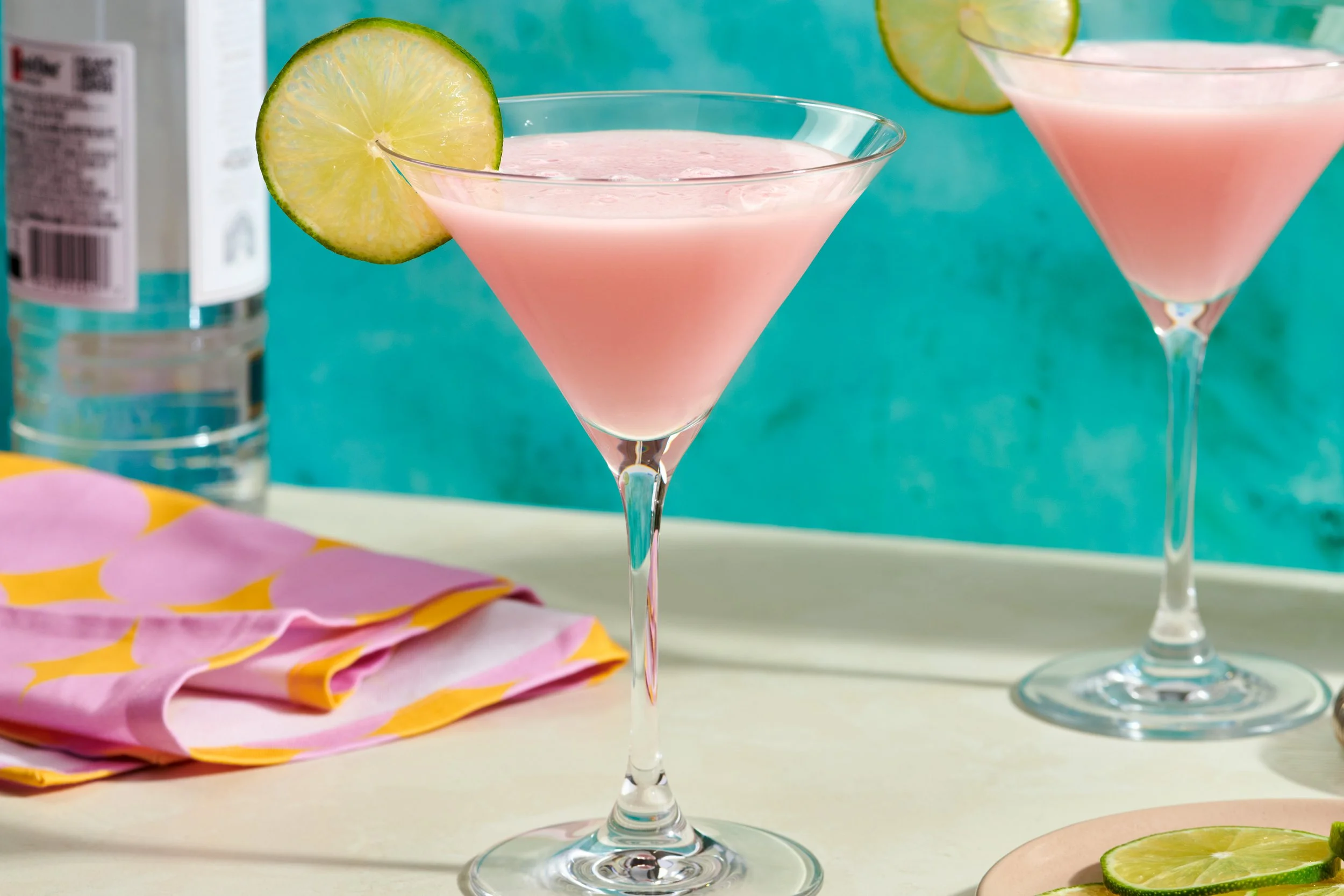 Two pink cocktails in martini glasses garnished with lime slices, with a blurred bottle in the background and colorful napkins on a white surface.