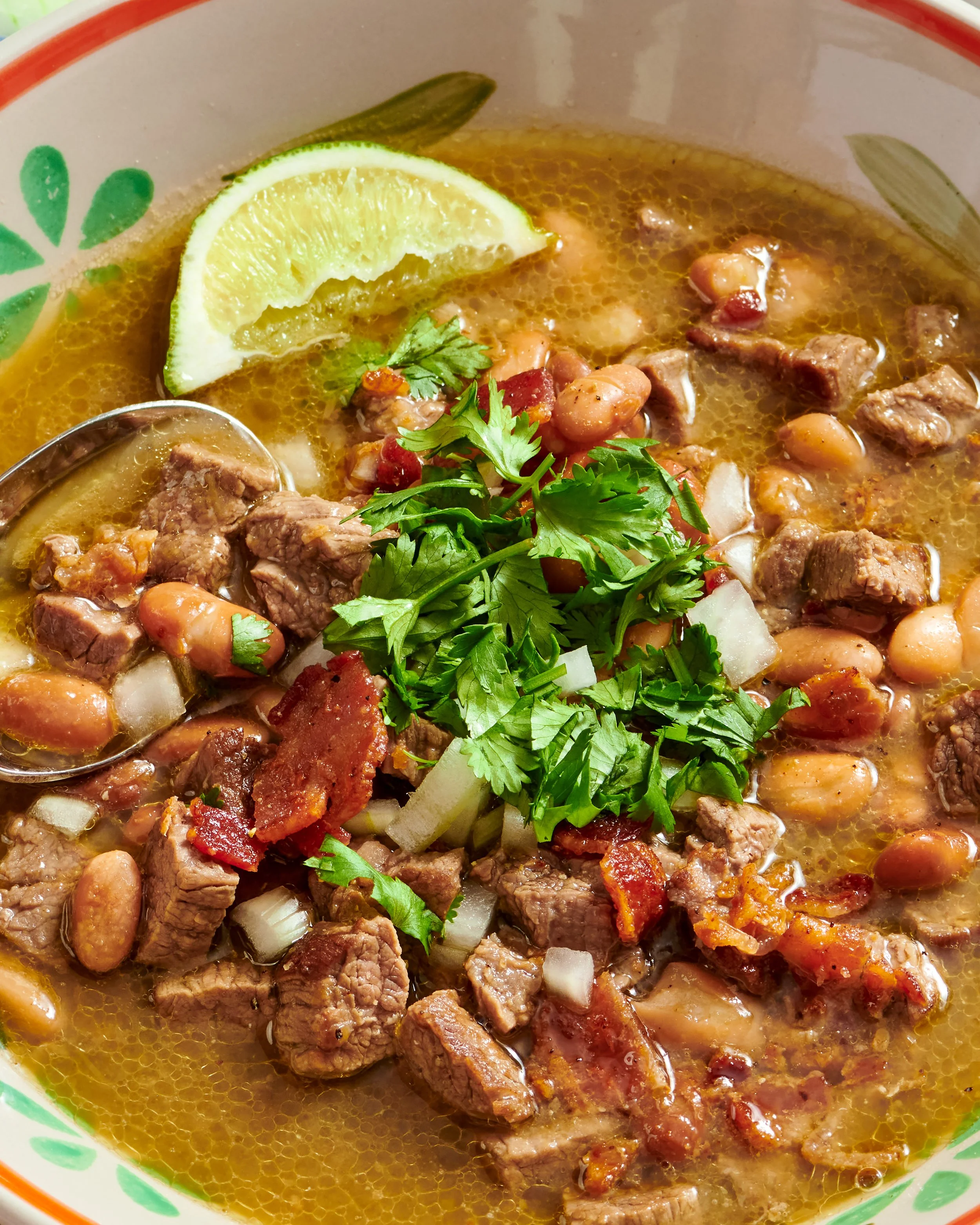 A bowl of beef stew with beans, cilantro, onion, a lemon wedge, and a spoon.