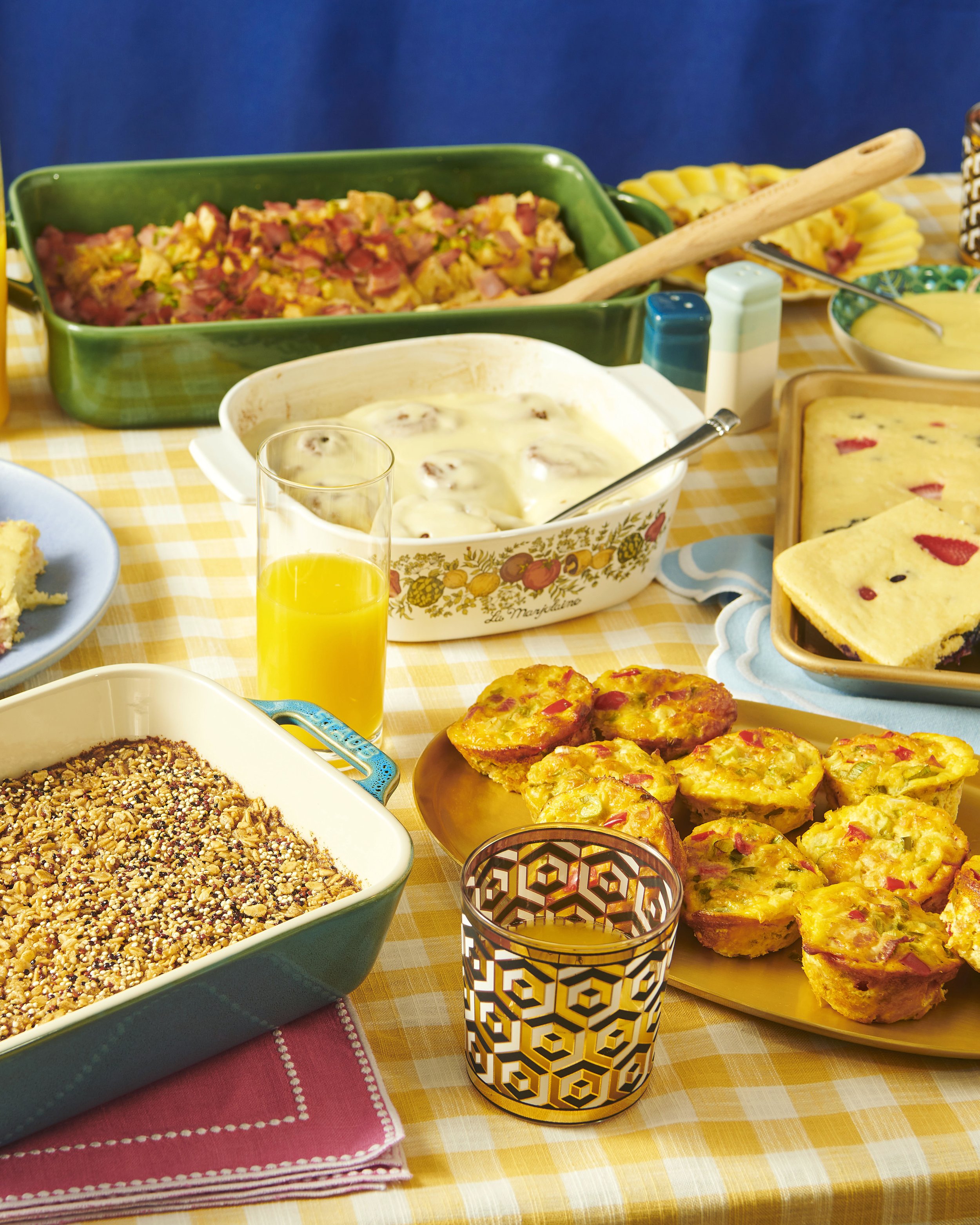 A table with a yellow checkered tablecloth featuring various breakfast foods, including a green casserole dish with scrambled eggs and ham, a white bowl with yogurt and fruit, a tray of mini quiches with vegetables and ham, and a glass of orange juic