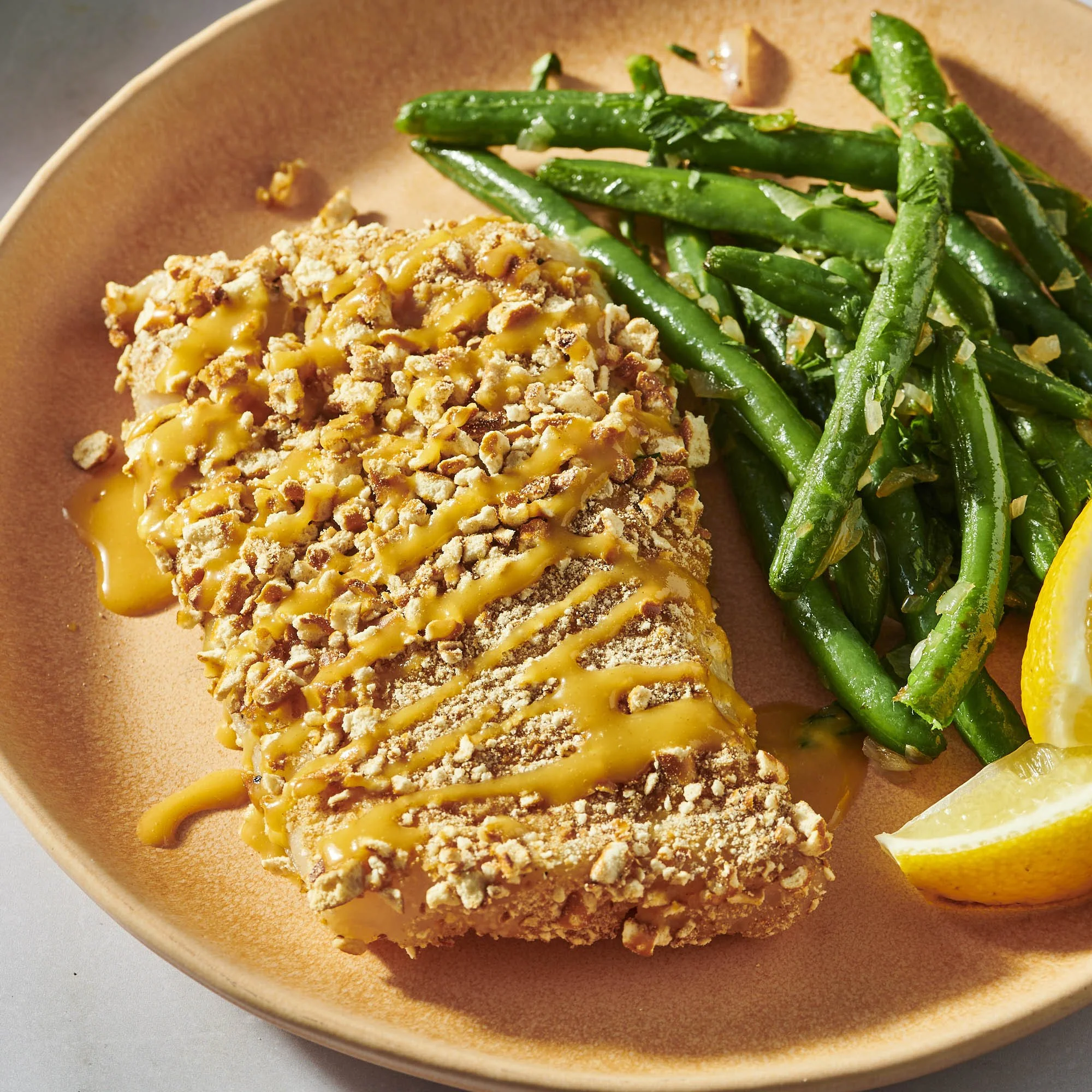 Close-up of a breaded fish fillet topped with a yellow sauce, served with green beans and lemon wedges on a pink plate.