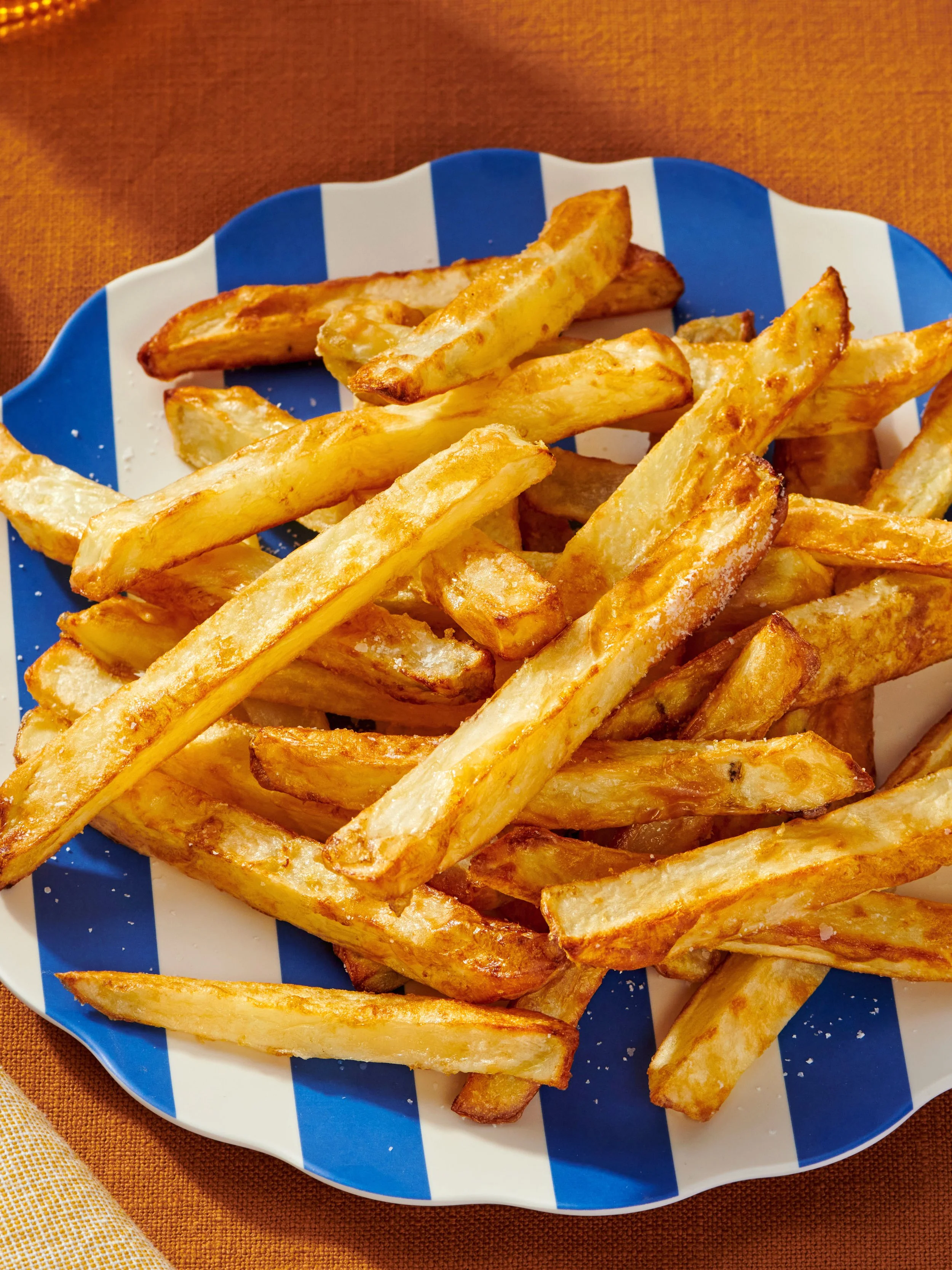 A plate of golden French fries on a white and blue striped dish, placed on an orange tablecloth.