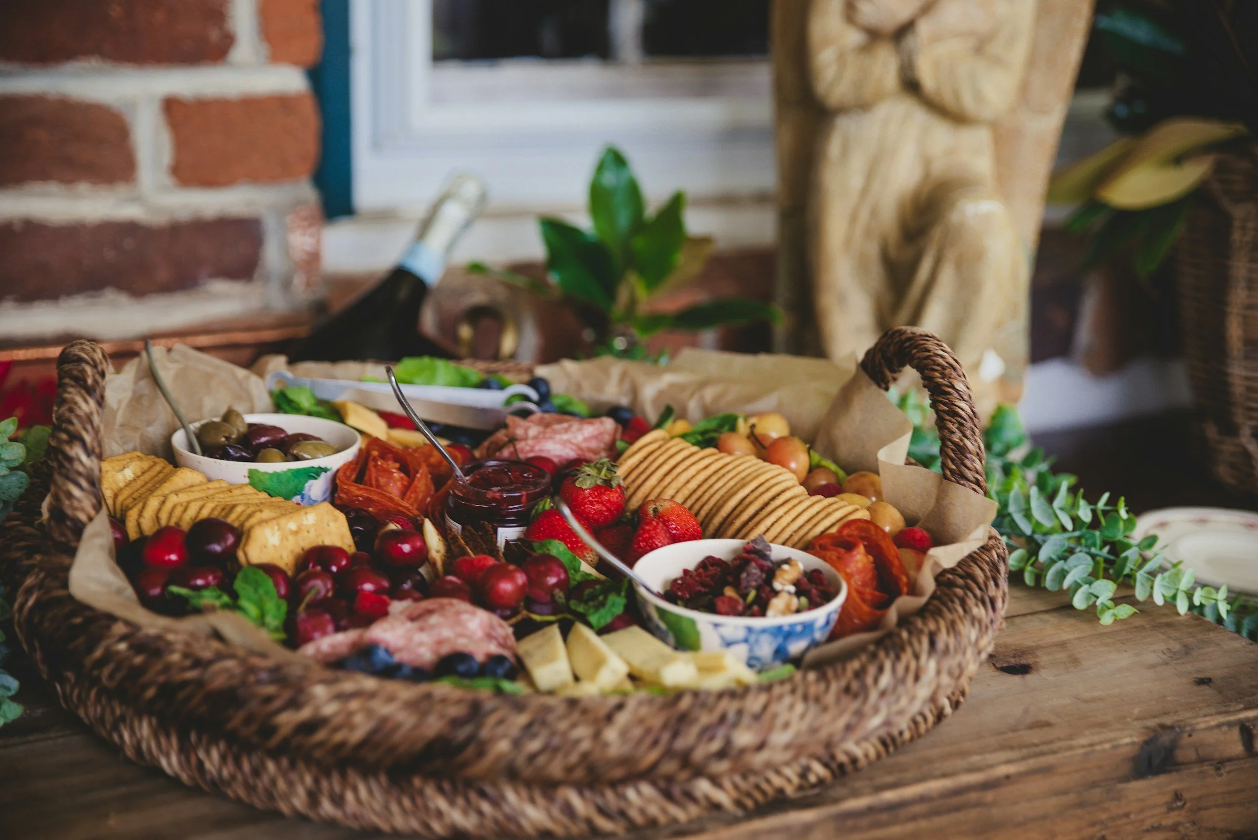 A woven basket filled with assorted fruits, crackers, and snacks on a wooden table, with a brick wall, window, and potted plants in the background.