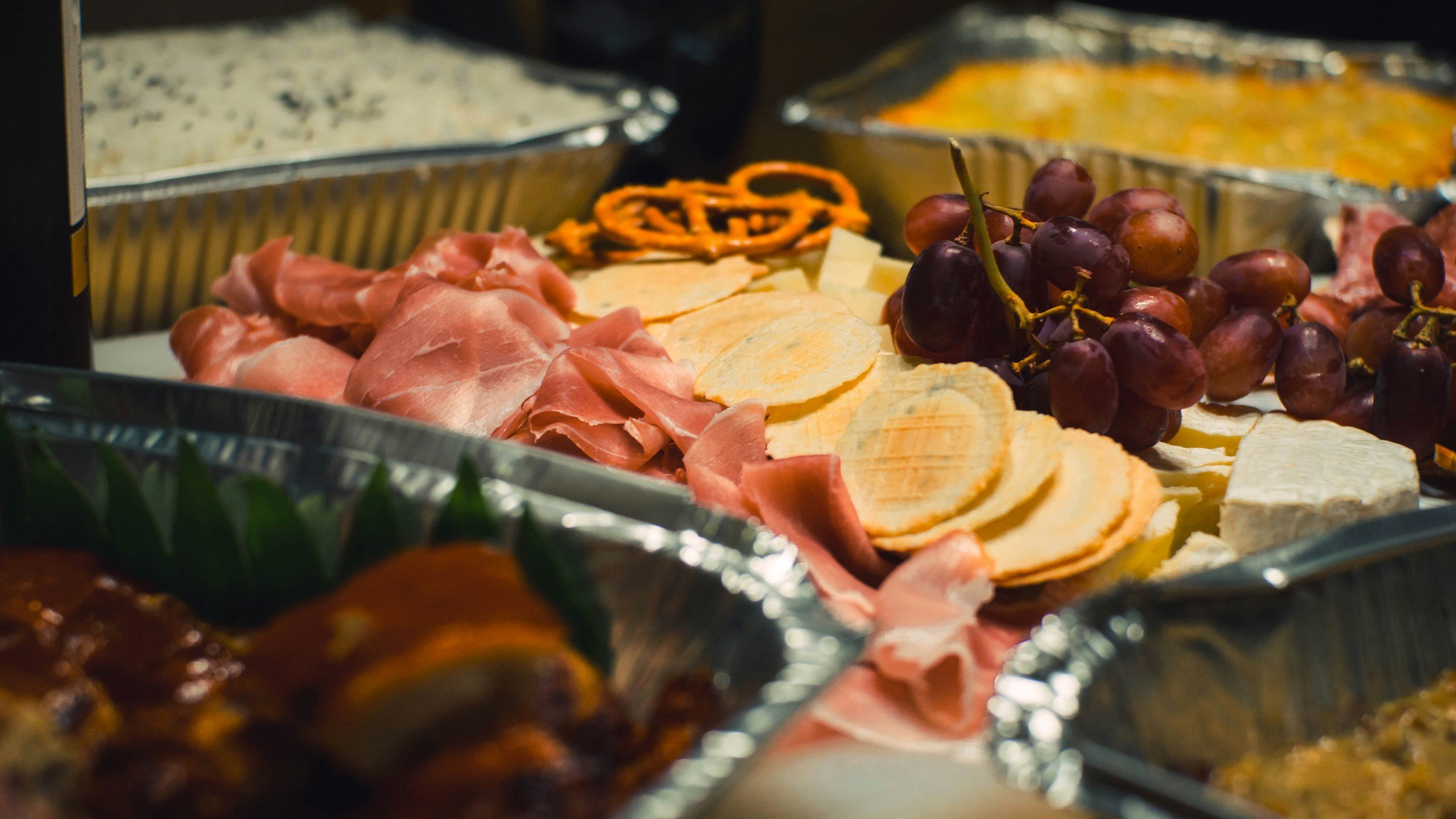 A buffet with various food items including grapes, sliced cheese, sliced ham, and other dishes in aluminum trays.