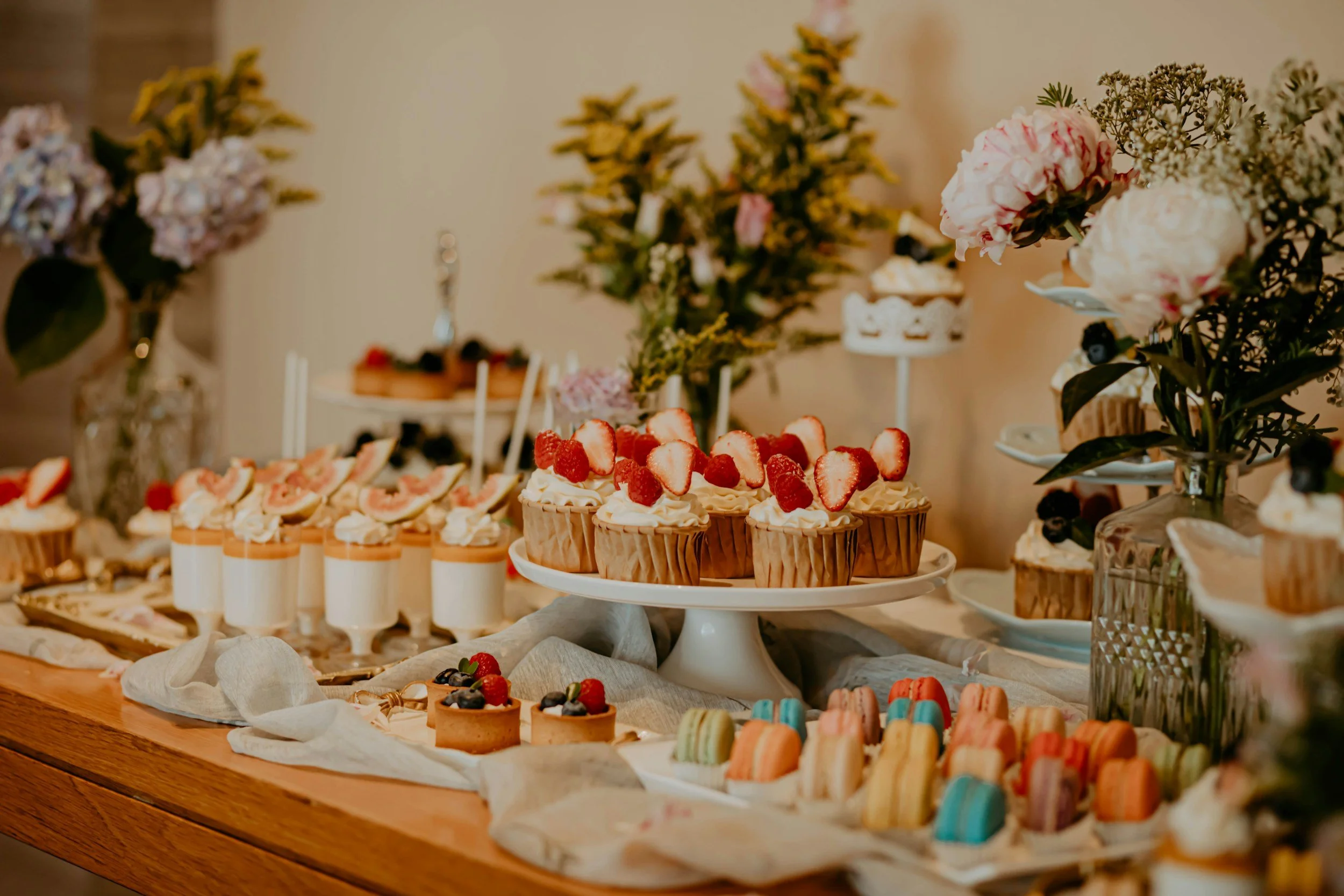Dessert table with various colorful pastries, cakes, and cookies decorated with strawberries and berries, surrounded by floral arrangements.
