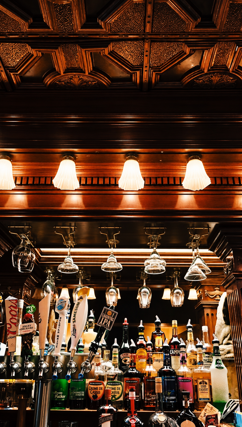 View of a bar with hanging glasses and a variety of liquor bottles, illuminated by warm light fixtures with a decorative wooden ceiling above.