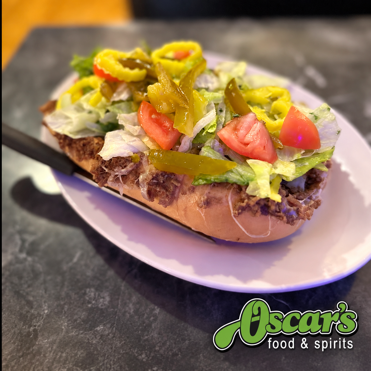 Open-faced burger sandwich topped with lettuce, tomato, pickles, and chopped peppers. The sandwich is on a bun with a beef patty and is served on a white plate. The bottom right corner shows a logo for Oscar's food & spirits.