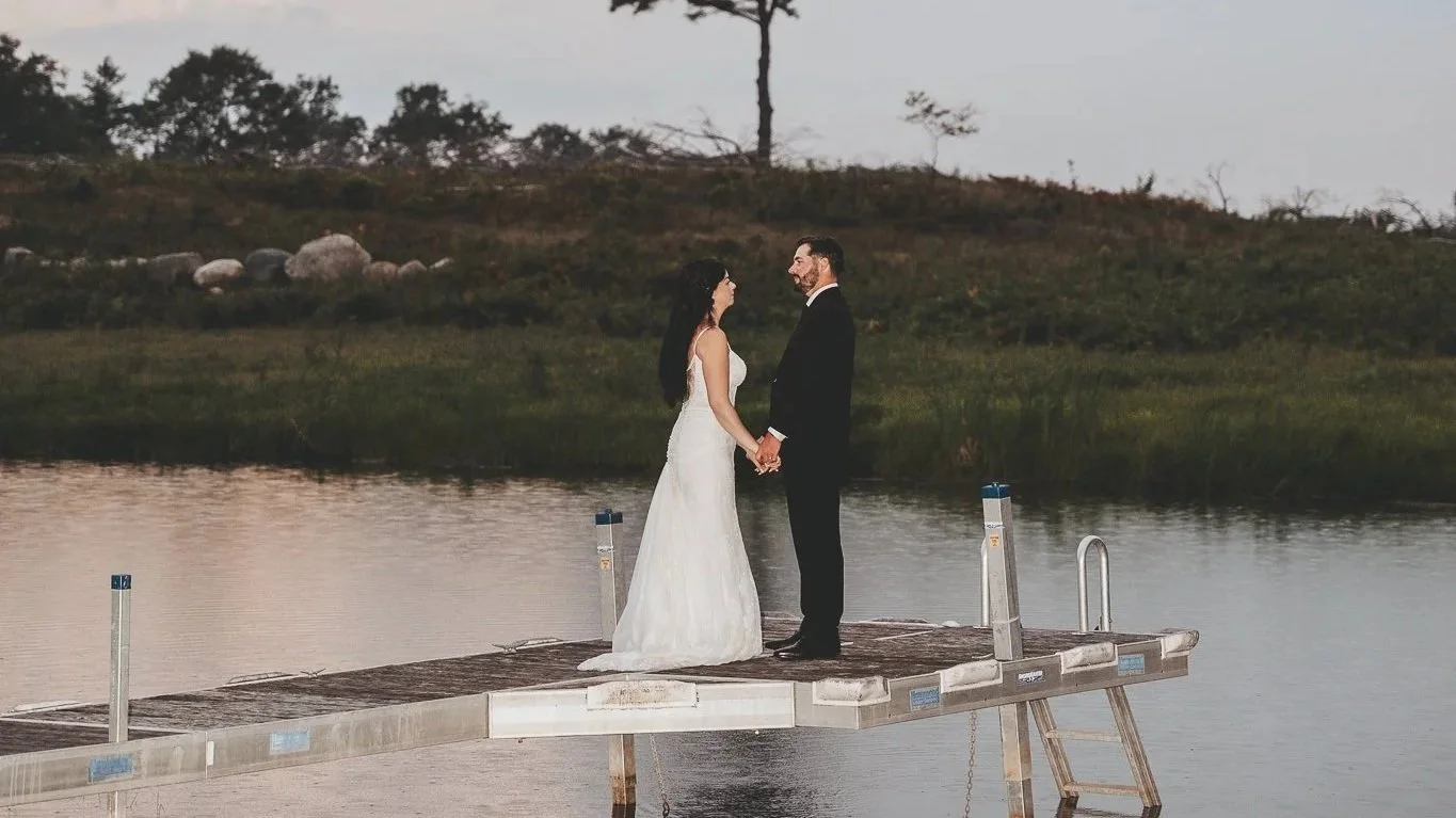 A bride and groom standing on a dock holding hands, facing each other, near a body of water with trees and rocks in the background.