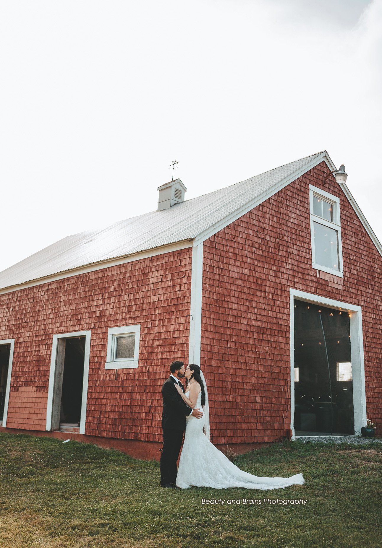 A bride and groom standing close together outside a red barn during a wedding, with the bride wearing a white gown and the groom in a black suit, in front of the barn with a grassy area.
