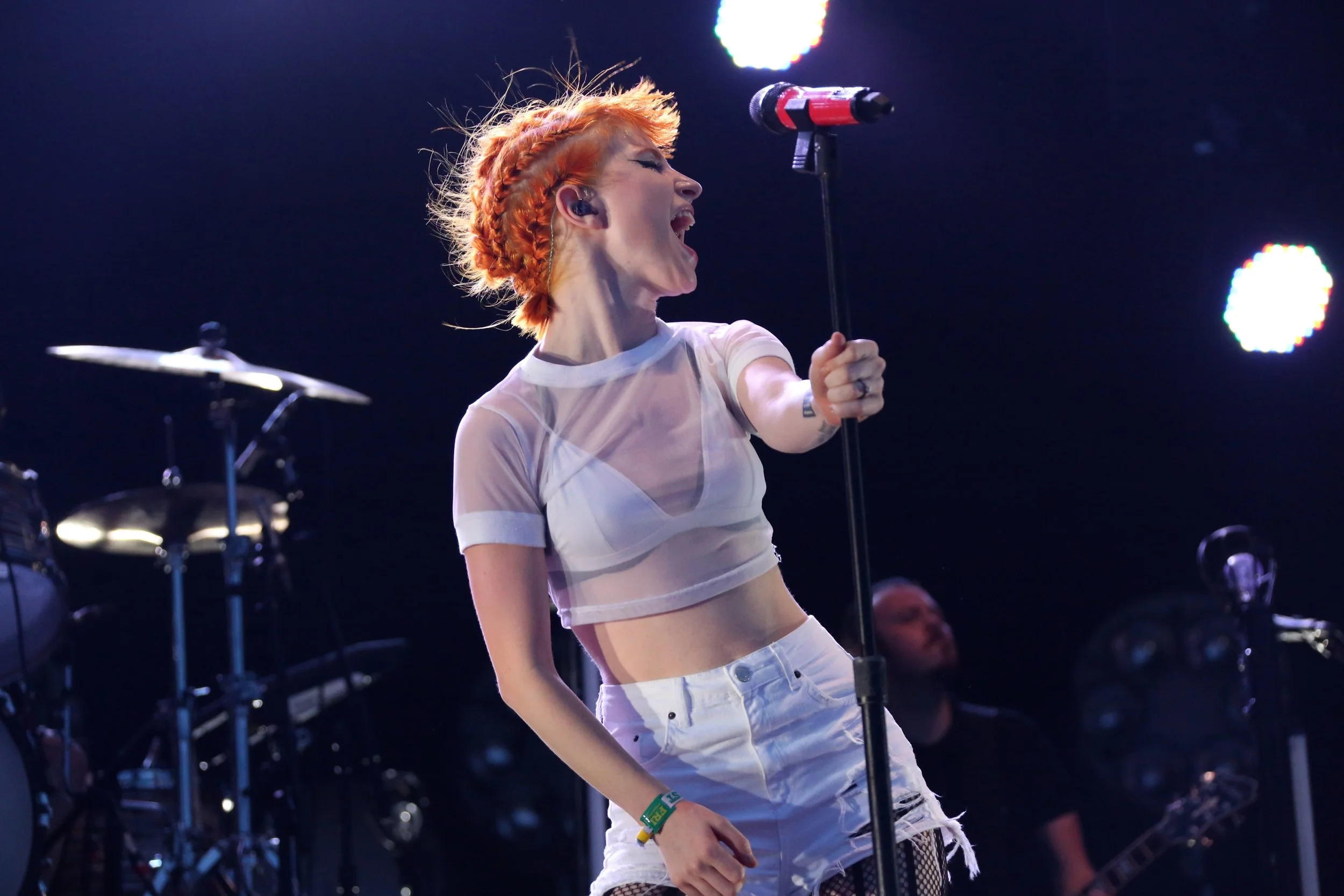 Hayley Williams with orange hair singing passionately on stage, holding a microphone, wearing a sheer white crop top and ripped white jeans, with stage lights in the background.
