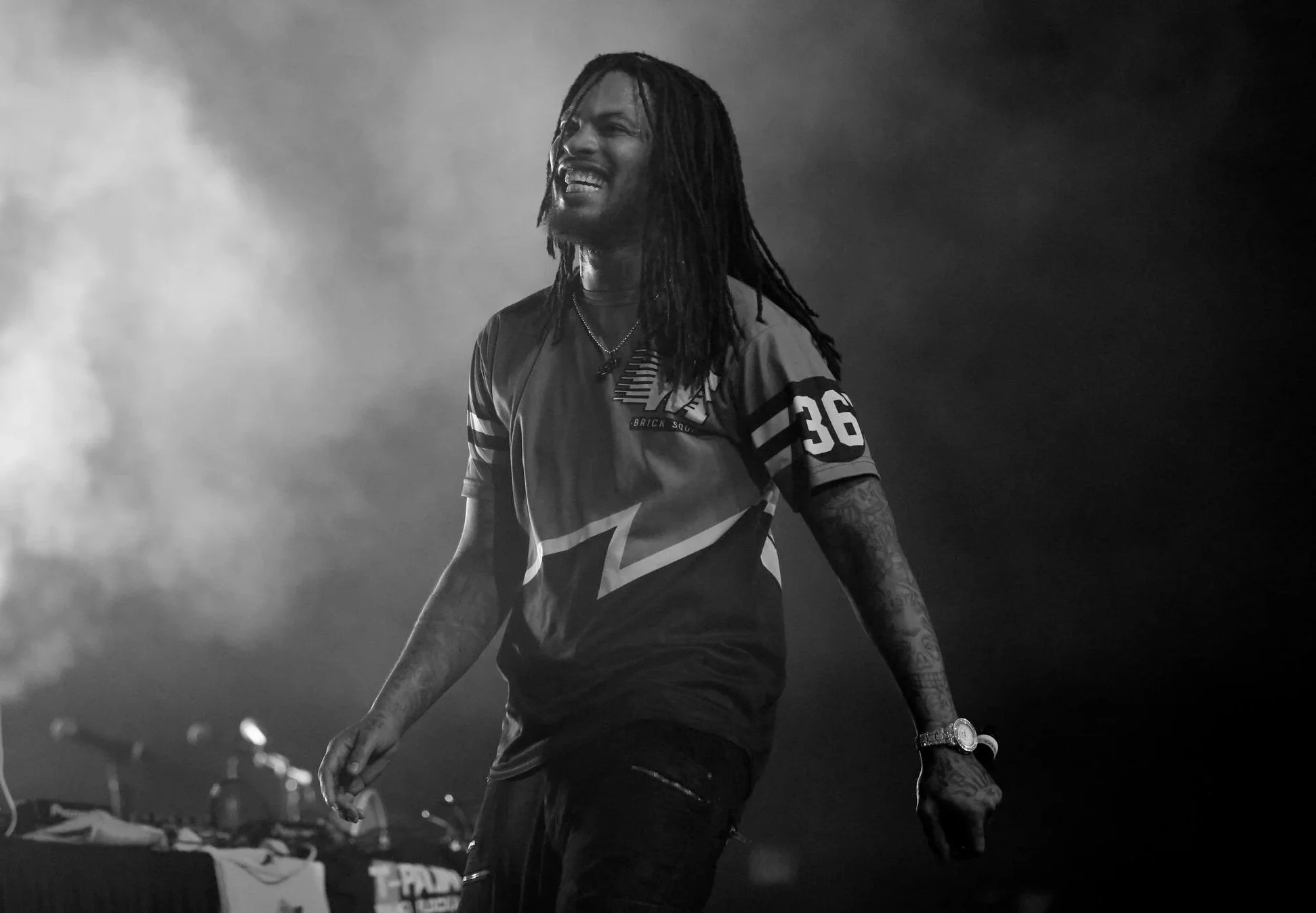 Waka Flocka Flame with long dreadlocks, tattoos, and a watch, wearing a sports jersey with the number 36, is smiling and walking in a dark, smoky environment, possibly during a concert or performance.