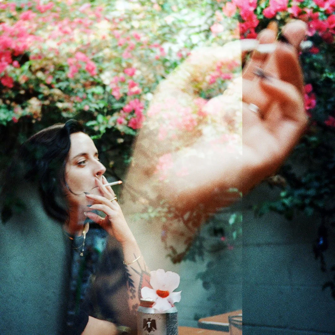 A woman with short dark hair and tattoos on her arm is smoking a cigarette while sitting at a table outdoors among pink flowering bushes.