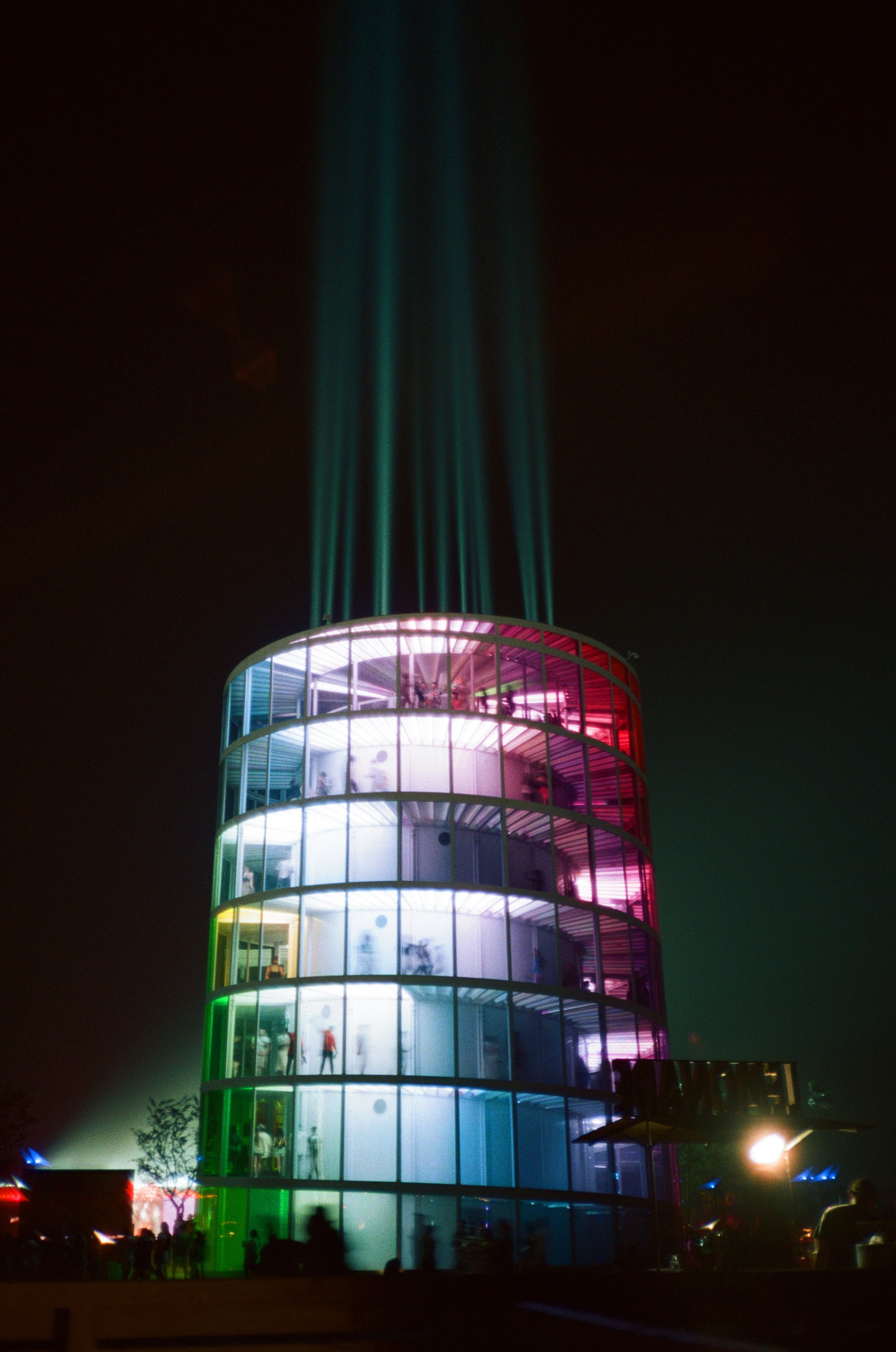 Nighttime view of a multi-story circular building at Coachella Music Festival with glass walls illuminated by multicolored lights, with people visible inside and outside, and beams of blue light shooting upwards into the dark sky.