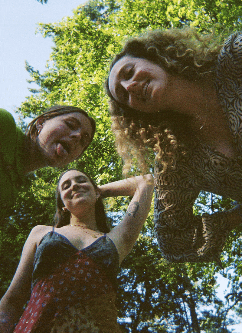 Three young women standing outside in a wooded area, looking down at the camera with smiles, surrounded by green trees and blue sky.