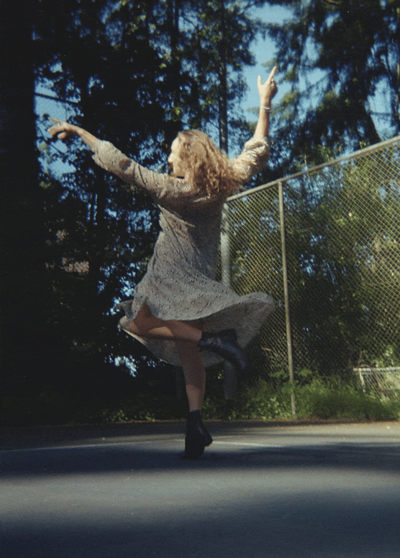 A woman with curly hair wearing a patterned dress and black boots dancing outdoors near a chain-link fence with trees in the background.