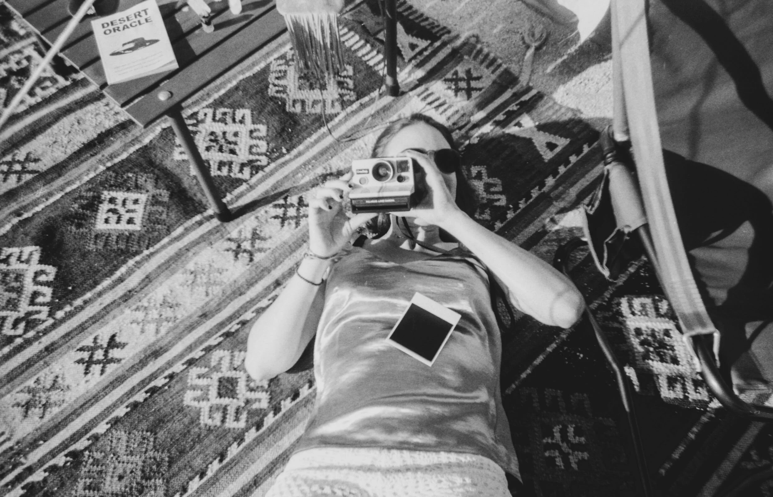 A person lying on a patterned rug taking a picture with a vintage camera, with a Polaroid camera and a smartphone nearby, and a book titled 'Desert Oracle' on a bench.