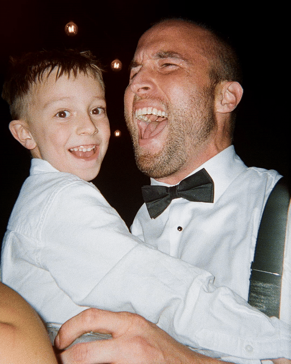 A man and a young boy are smiling and embracing at a celebration, both dressed formally with the man wearing a tuxedo and bow tie.