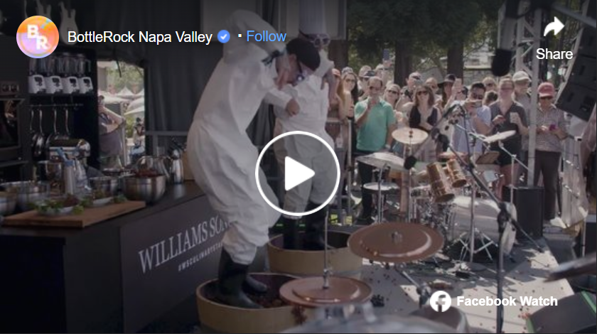 Musicians in white suits performing on stage at BottleRock Napa Valley music festival, with a crowd of concert-goers in the background.