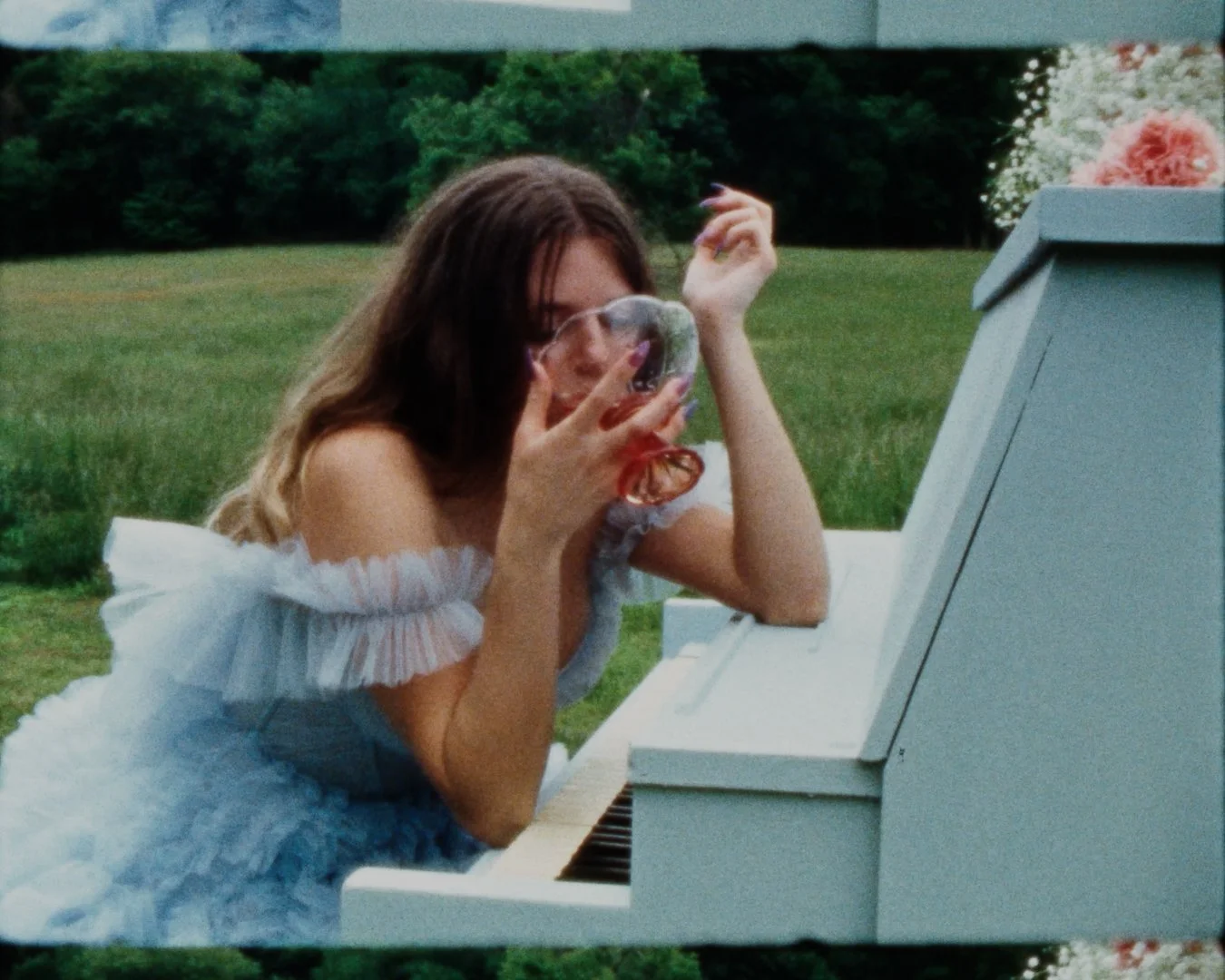 A young woman with brown hair wearing an off-shoulder ruffled white top playing a white upright piano outdoors on a grassy field with trees in the background, blowing bubbles.