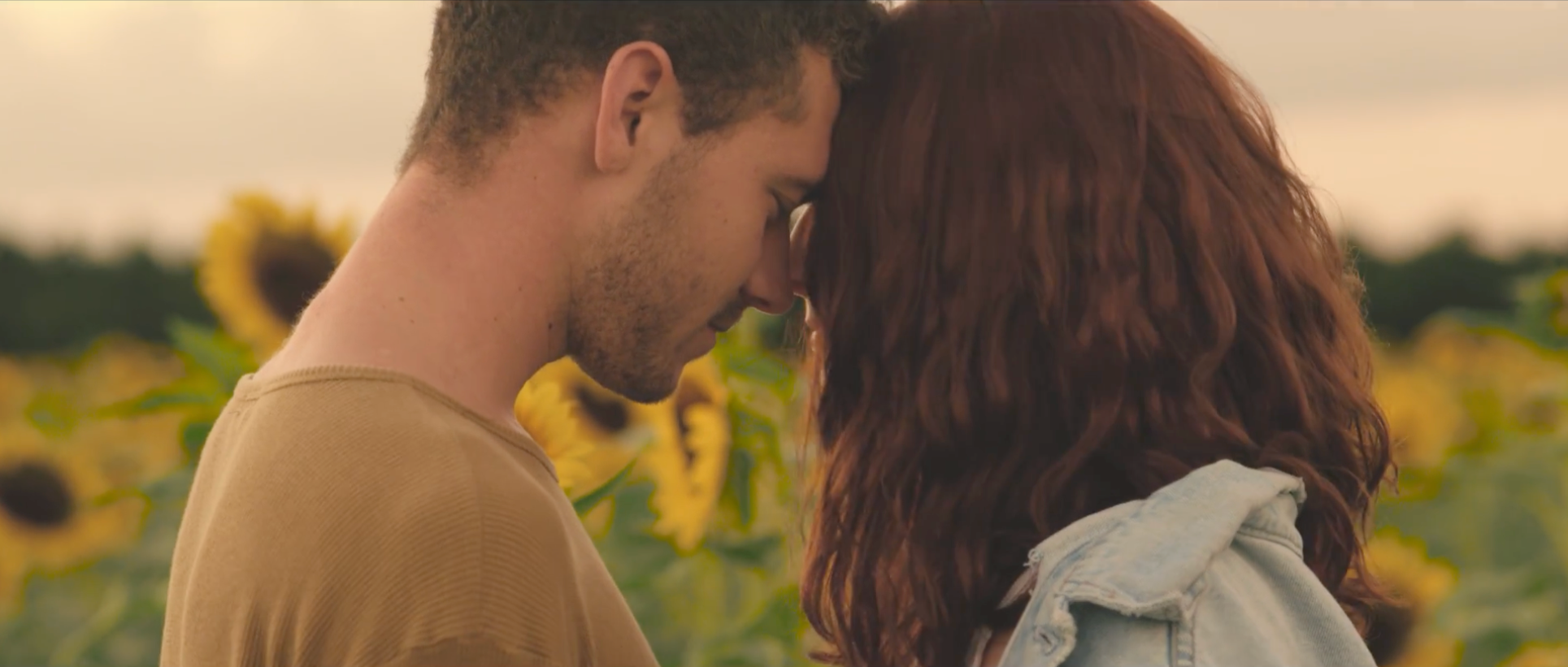 A young man and woman with their foreheads touching, standing close in a sunflower field during sunset.