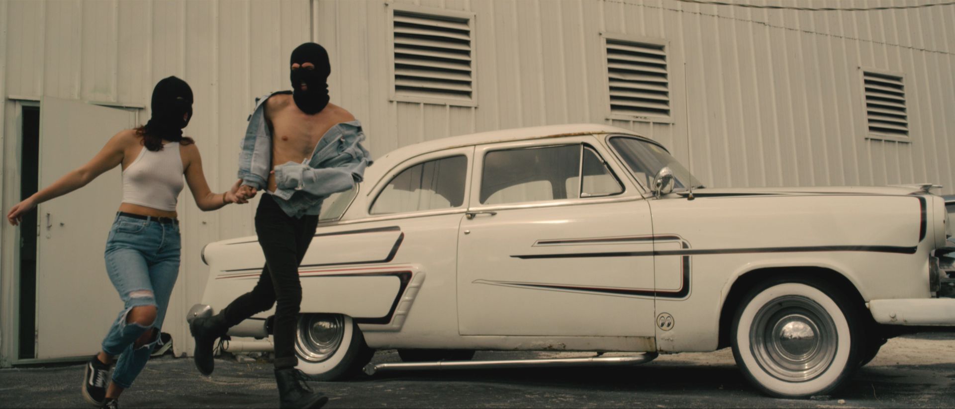 Two people wearing black ski masks and casual clothes are near a vintage white car with a black stripe, in front of a metal building.