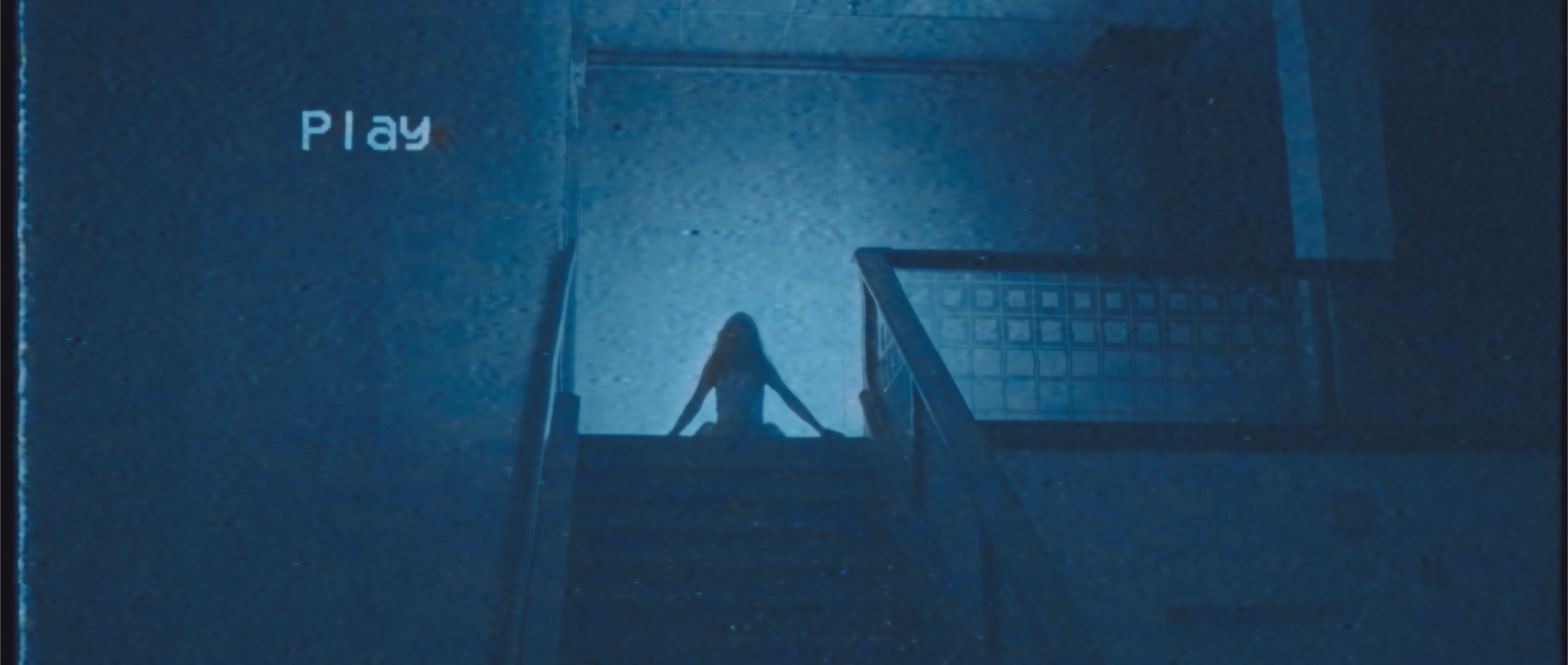 A person sitting on top of a staircase at night, with a large illuminated 'Play' sign on a wall nearby, and a metal gate at the bottom of the stairs.
